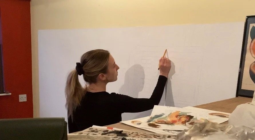 Artist sketching on a large white canvas with a table in front covered in used paint pallets.