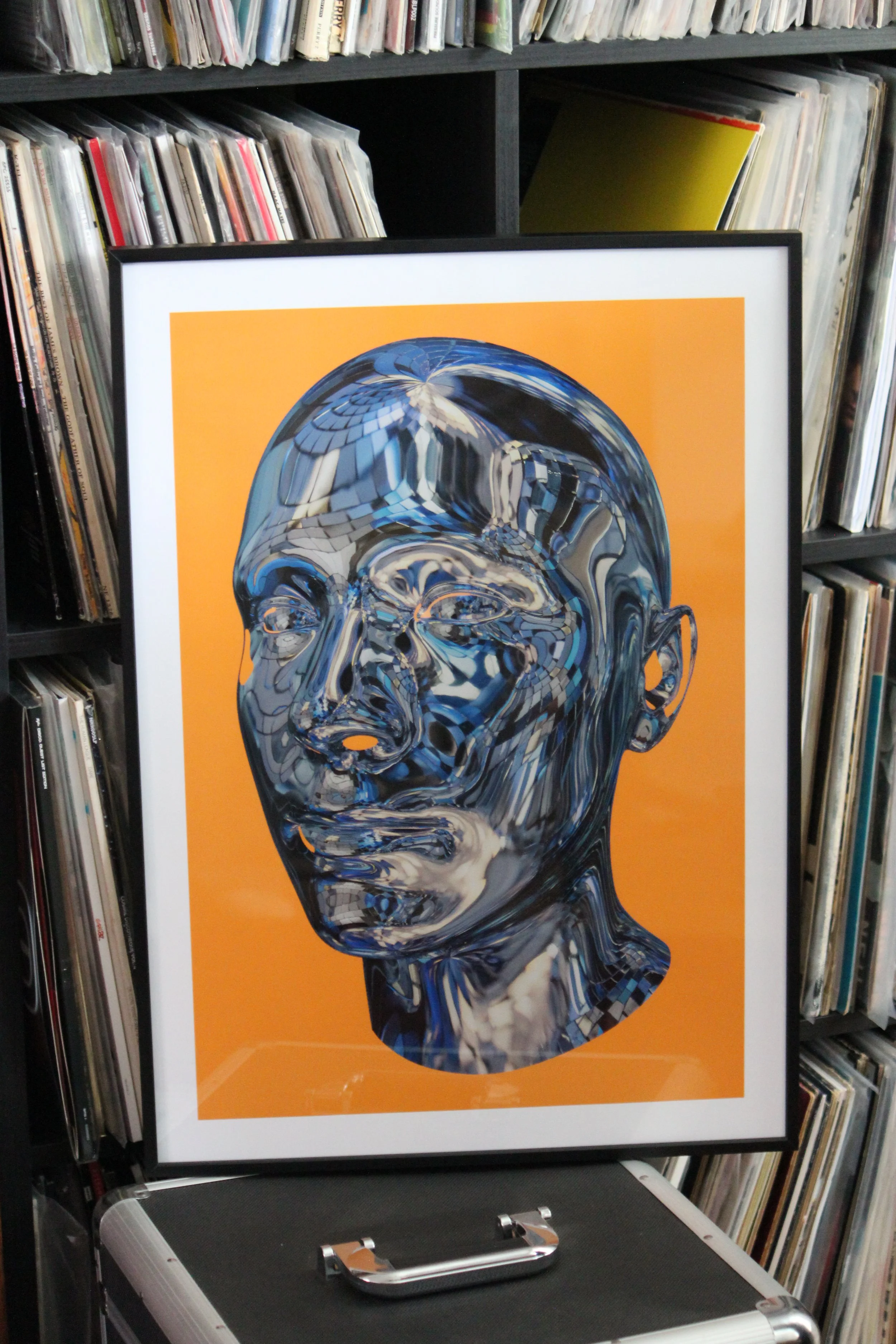 Framed art print of a male head made out of blue disco ball pieces on an orange background. Displayed on a bookshelf full of vinyl records.