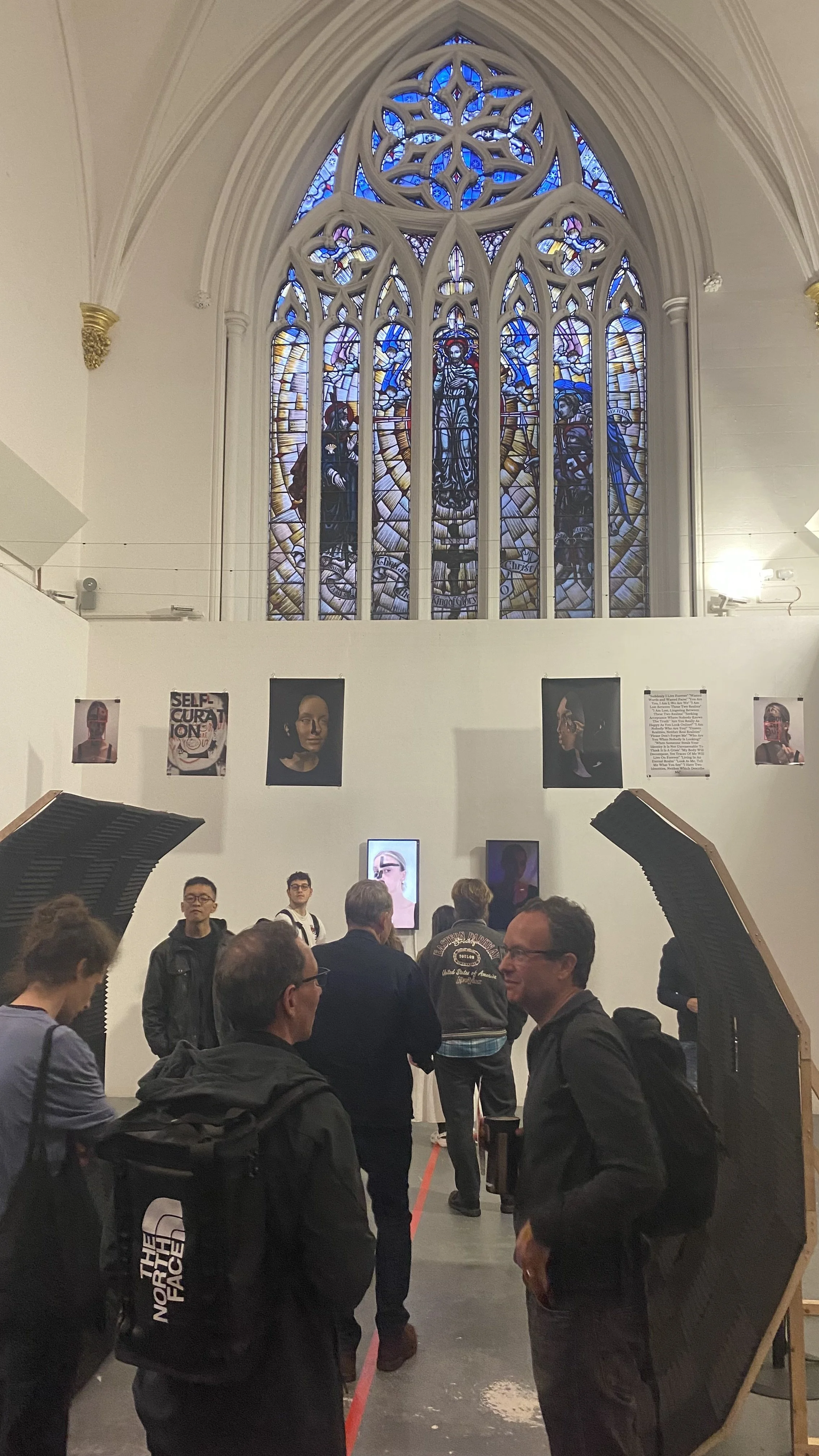 People gathered in an art gallery or museum space with a large stained glass window overhead, artworks displayed on the wall, and digital screens showing portraits or images related to the exhibition.