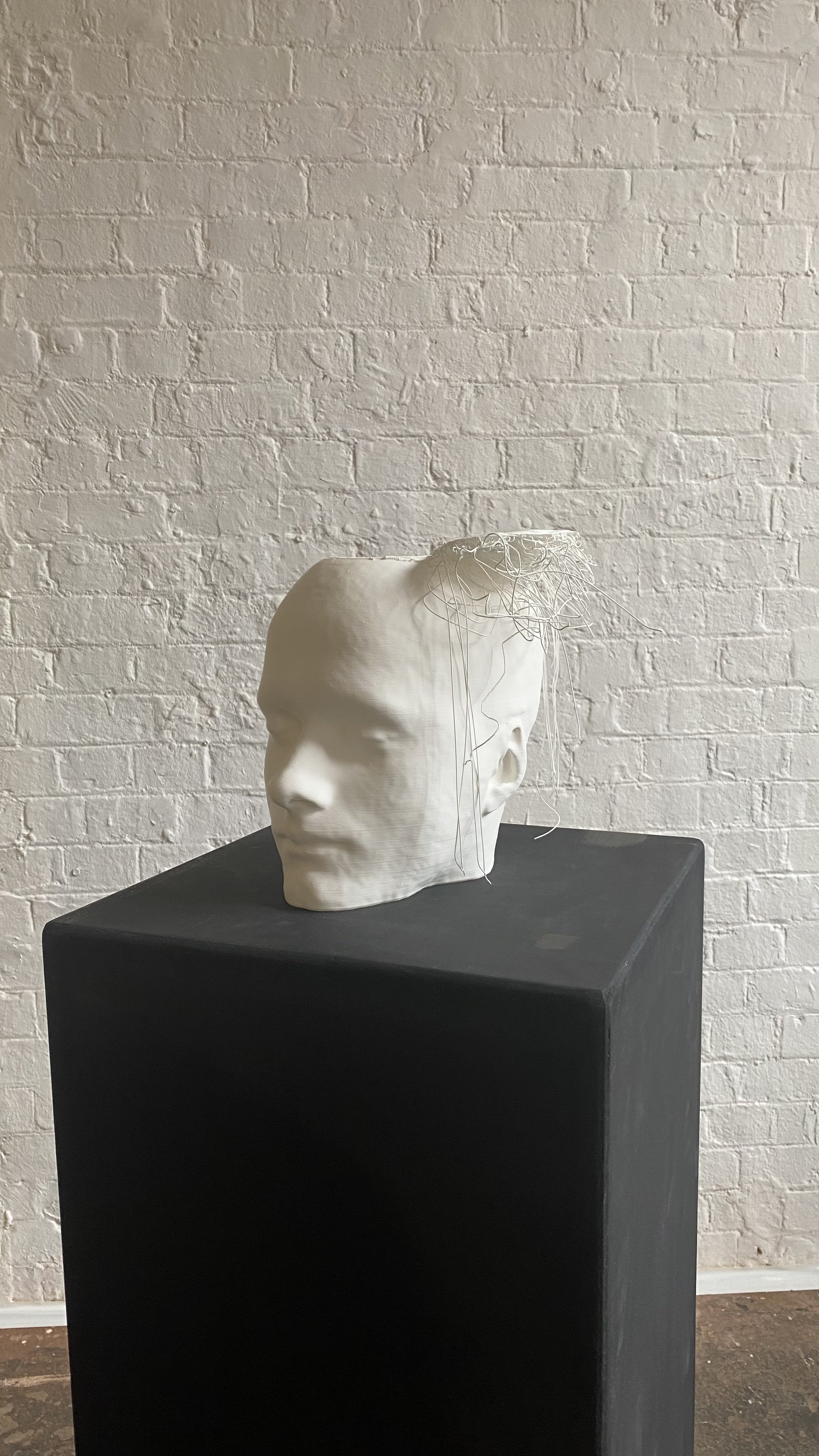 A white sculpture of a human head missing the top part to its head, falling apart and hanging down the side, displayed on a black plinth against a white brick wall.