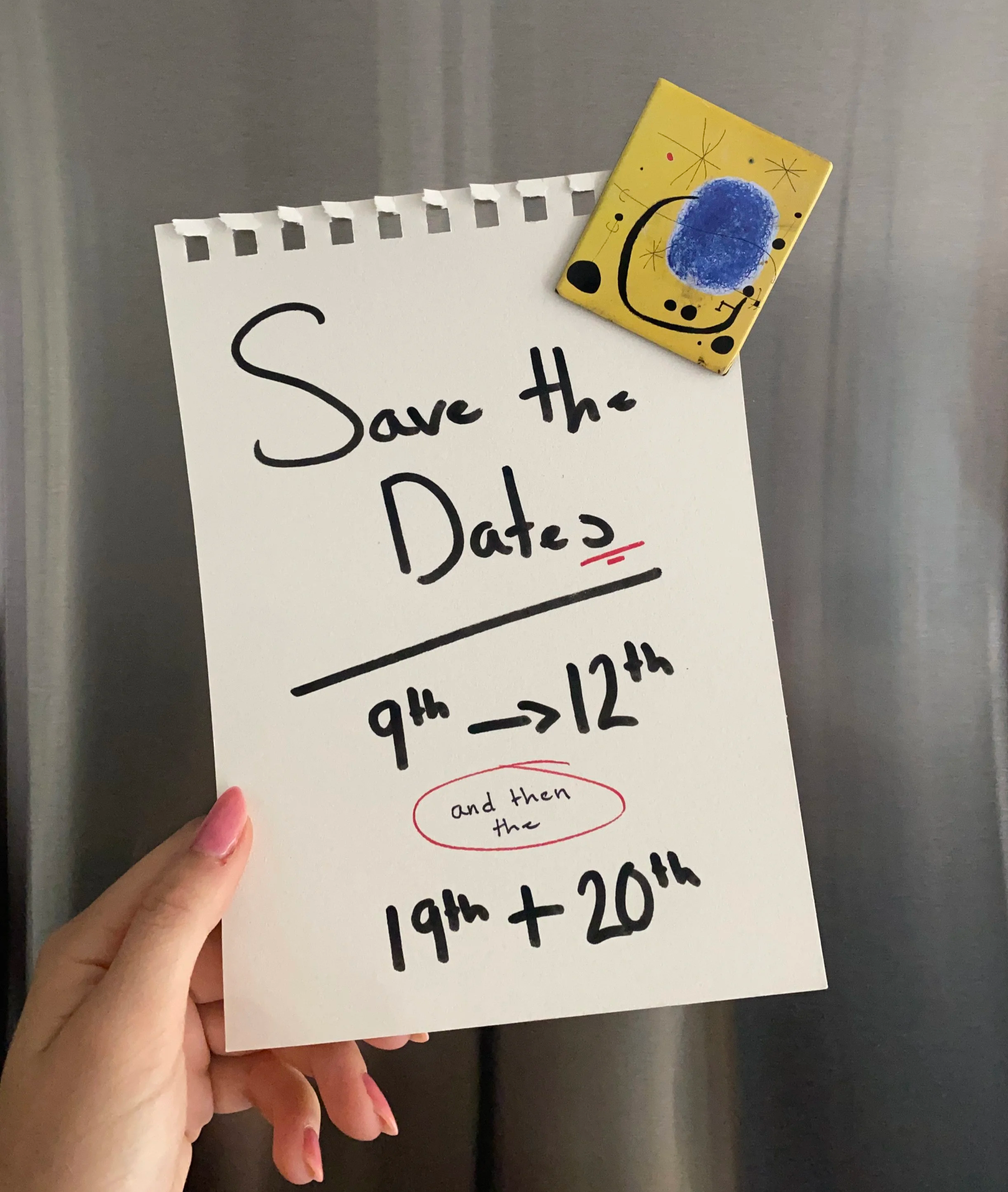 Hand placing a handwritten note on white paper with black and red markers, reading 'Save the Dates,' onto a stainless steel magnetic board. A yellow magnet is in the top right-hand corner.
