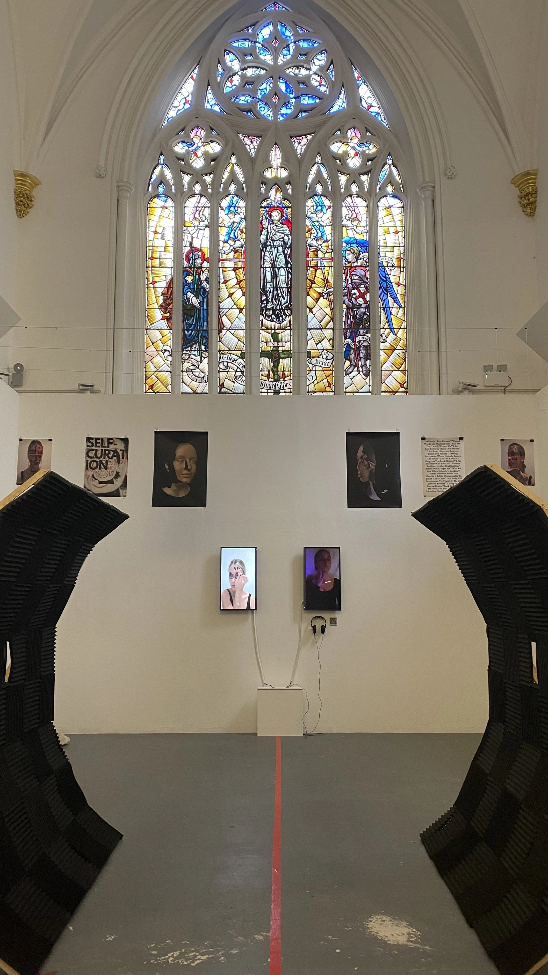 An art installation in a church showcases digital portraits on a white wall and prints suspended from the ceiling below a colourful stained glass window with religious depictions.