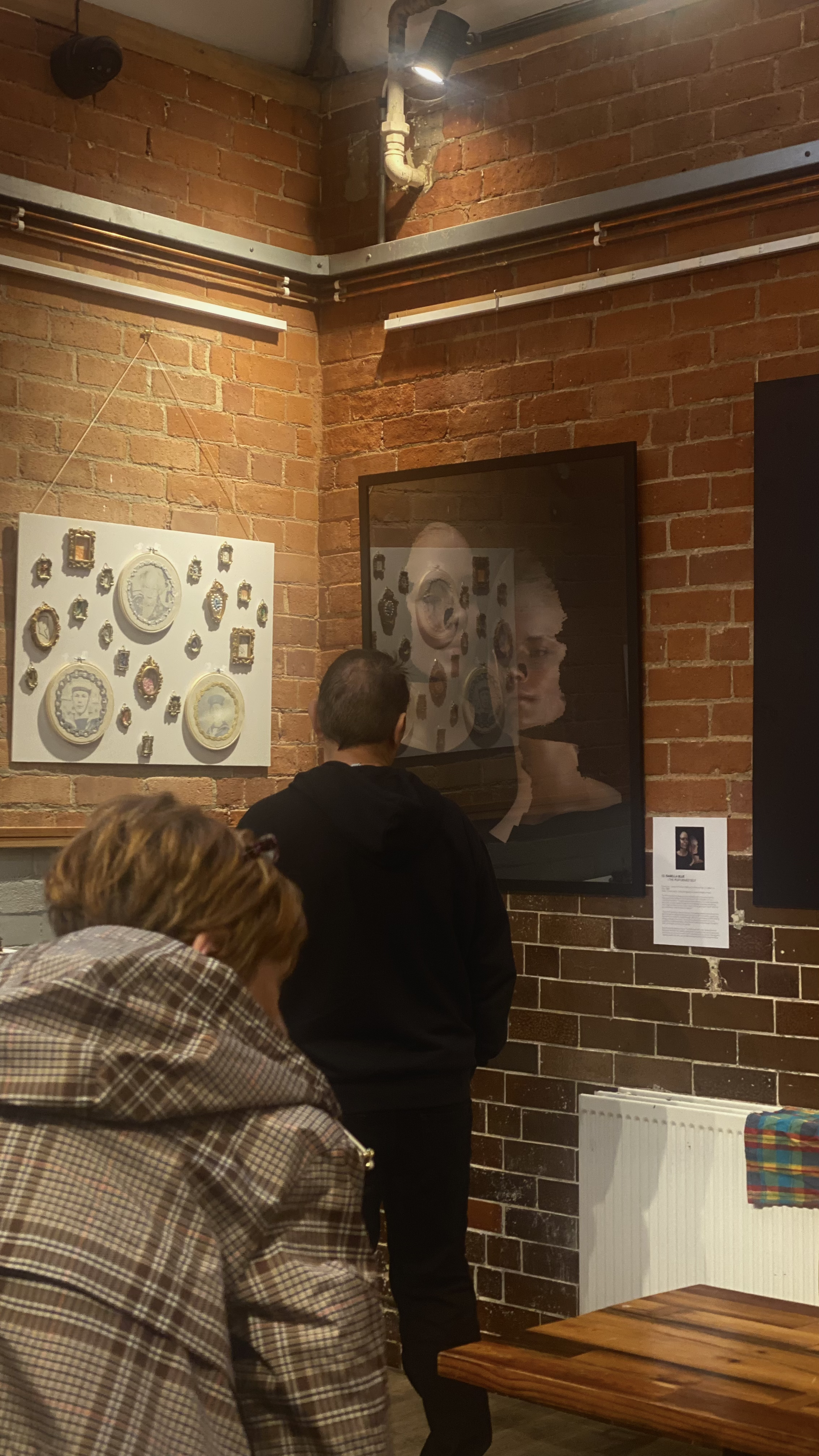 People viewing framed artwork in a gallery against red coloured brick walls.