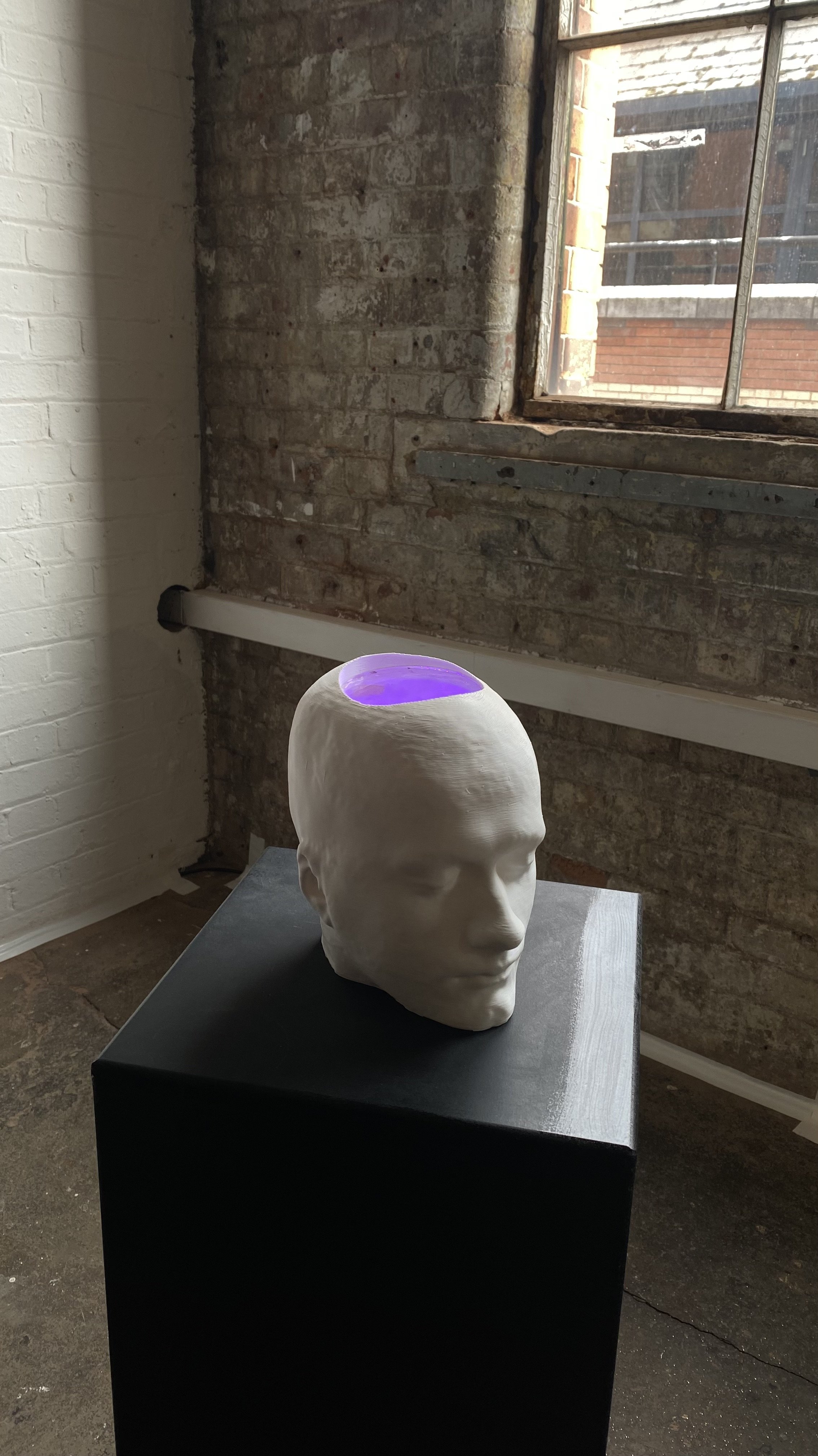A 3D printed head sculpture on a black plinth. The head is hollowed out inside and a light inside makes it glow purple.