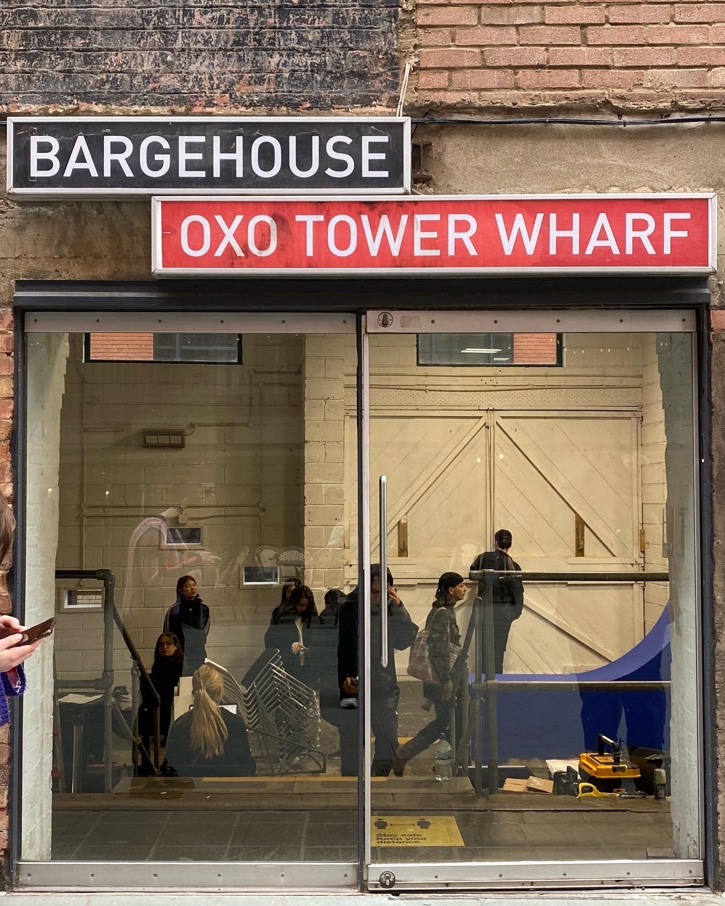 The main entrance to the gallery with the sign "Bargehouse, Oxo Tower Wharf" above. The doors are glass, showing individuals inside the building.