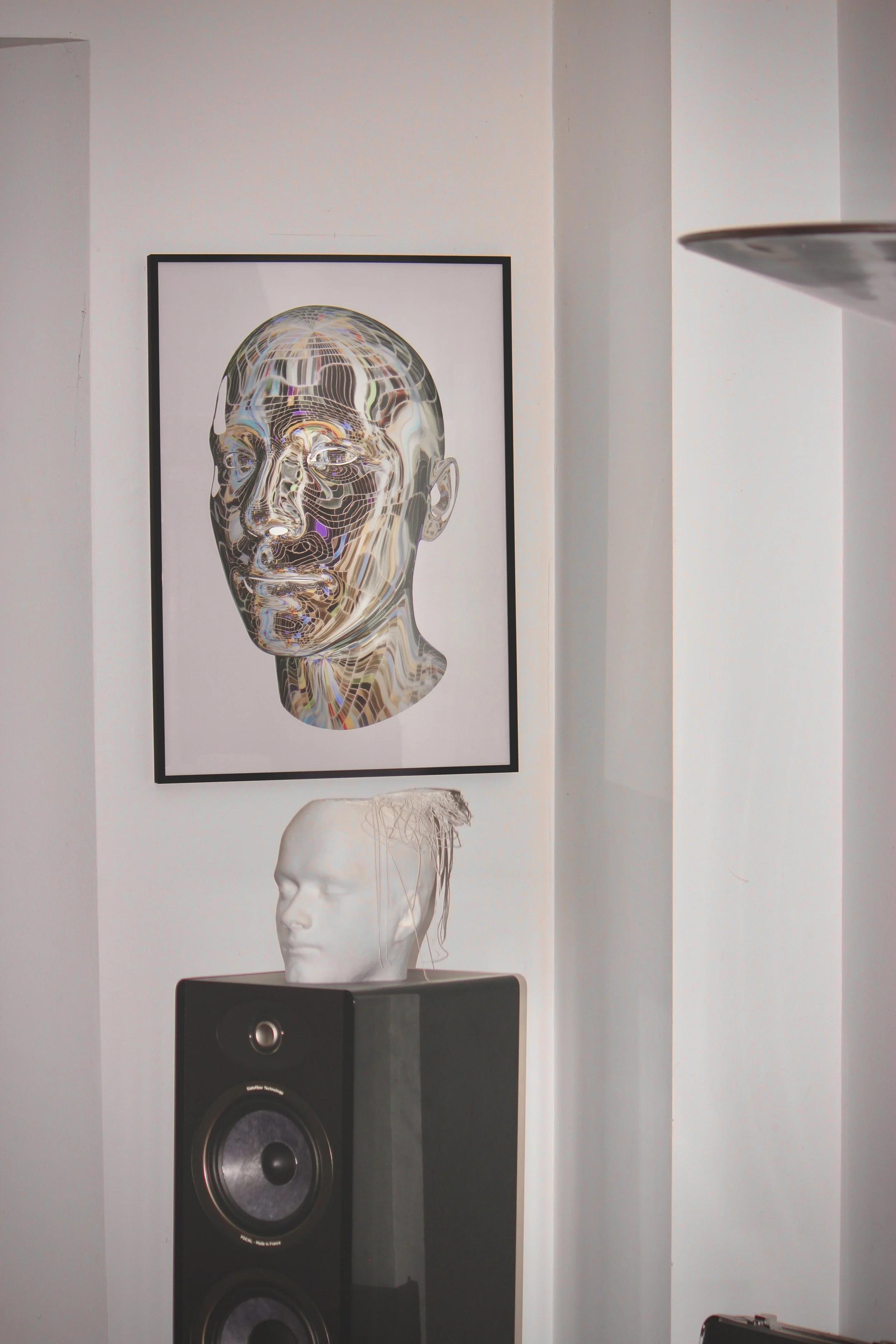 Art print of a male head made out of silver disco ball pieces on a white background, framed and hanging on a wall. A white sculpture of a head and a black speaker are below the artwork.