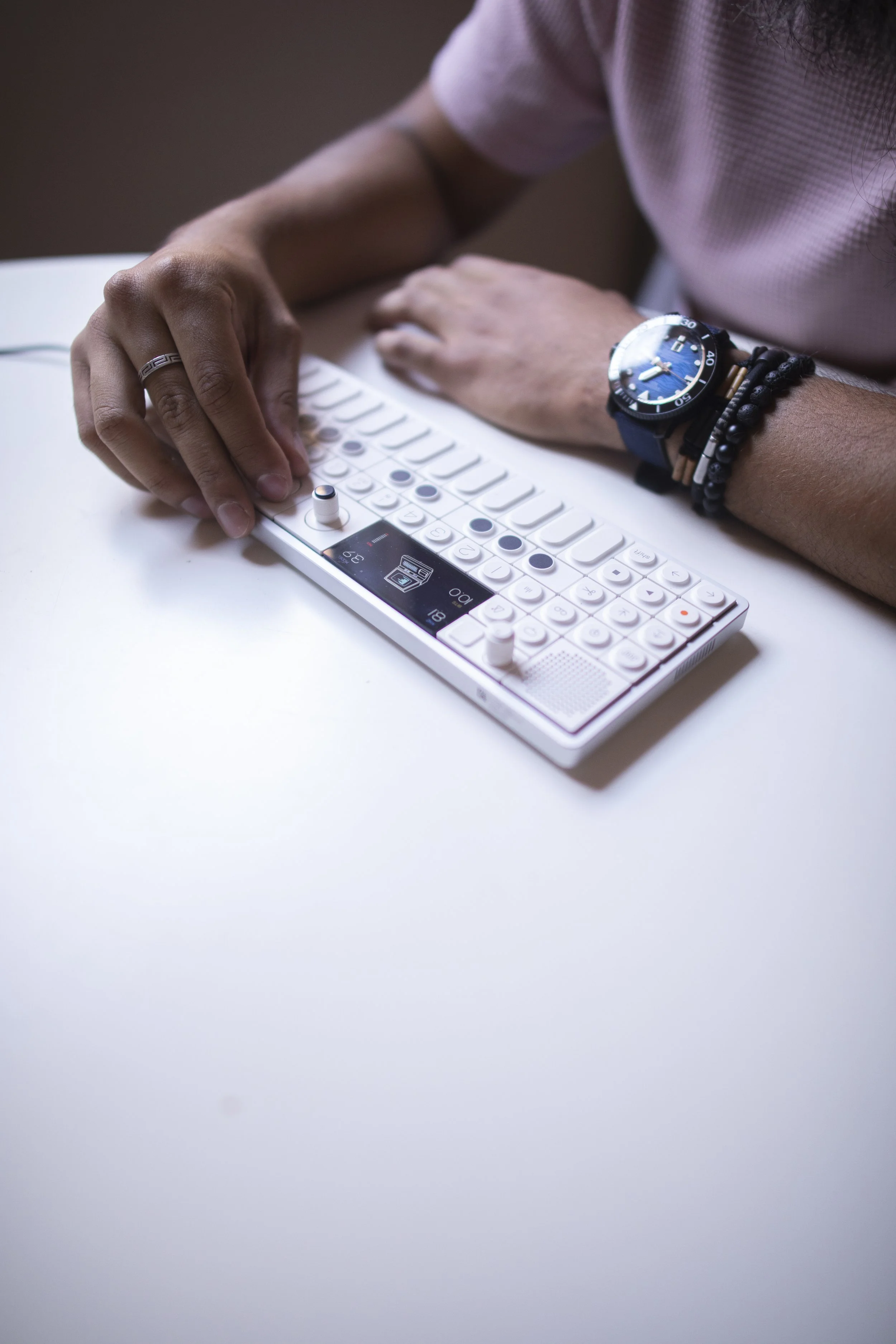 Persona tocando un secuenciador musical portátil en una mesa blanca, usando varias pulseras y una camiseta de manga corta rosada.