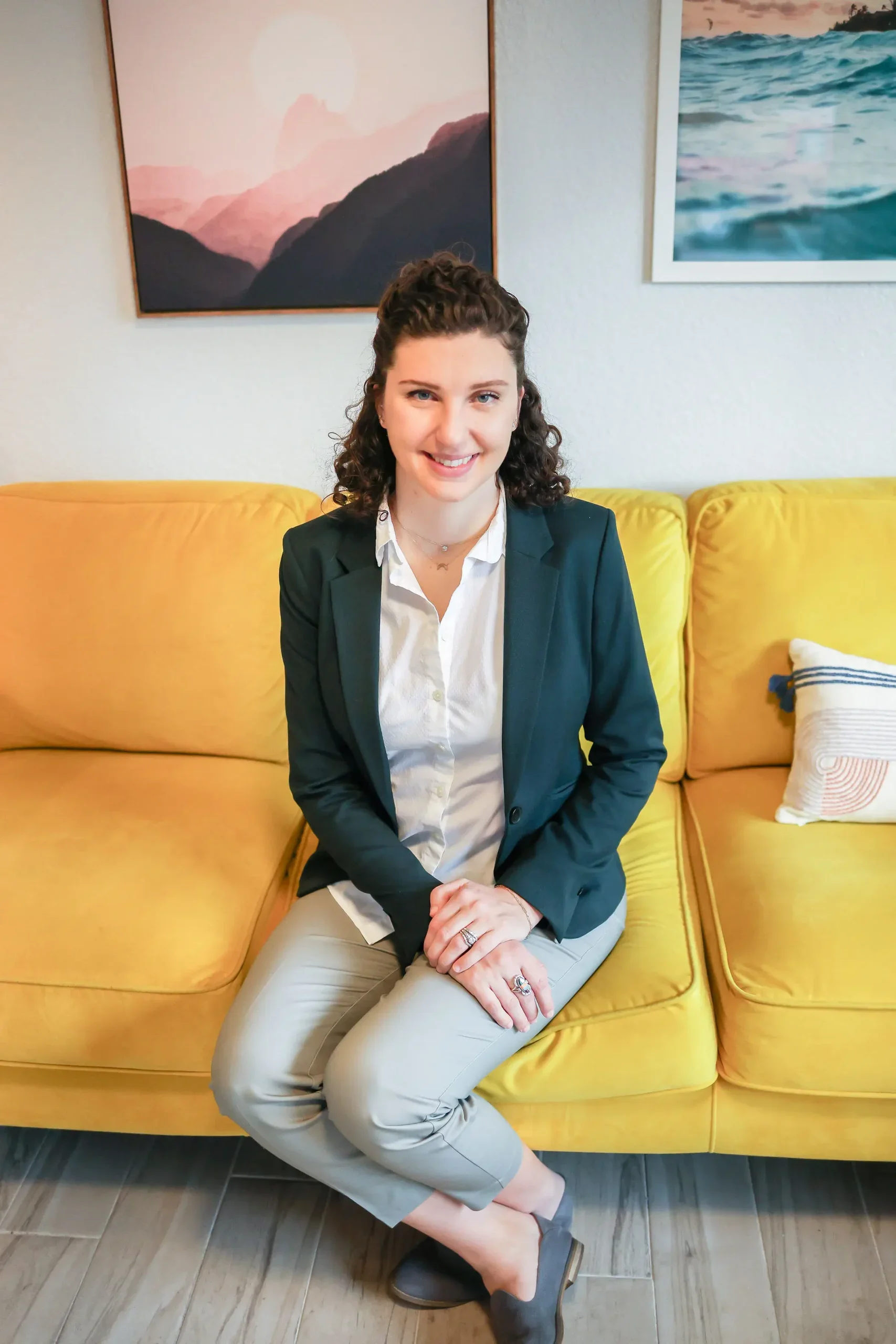 A woman with curly hair sitting on a yellow couch, smiling at the camera, in a room with framed wall art and decorative pillows.