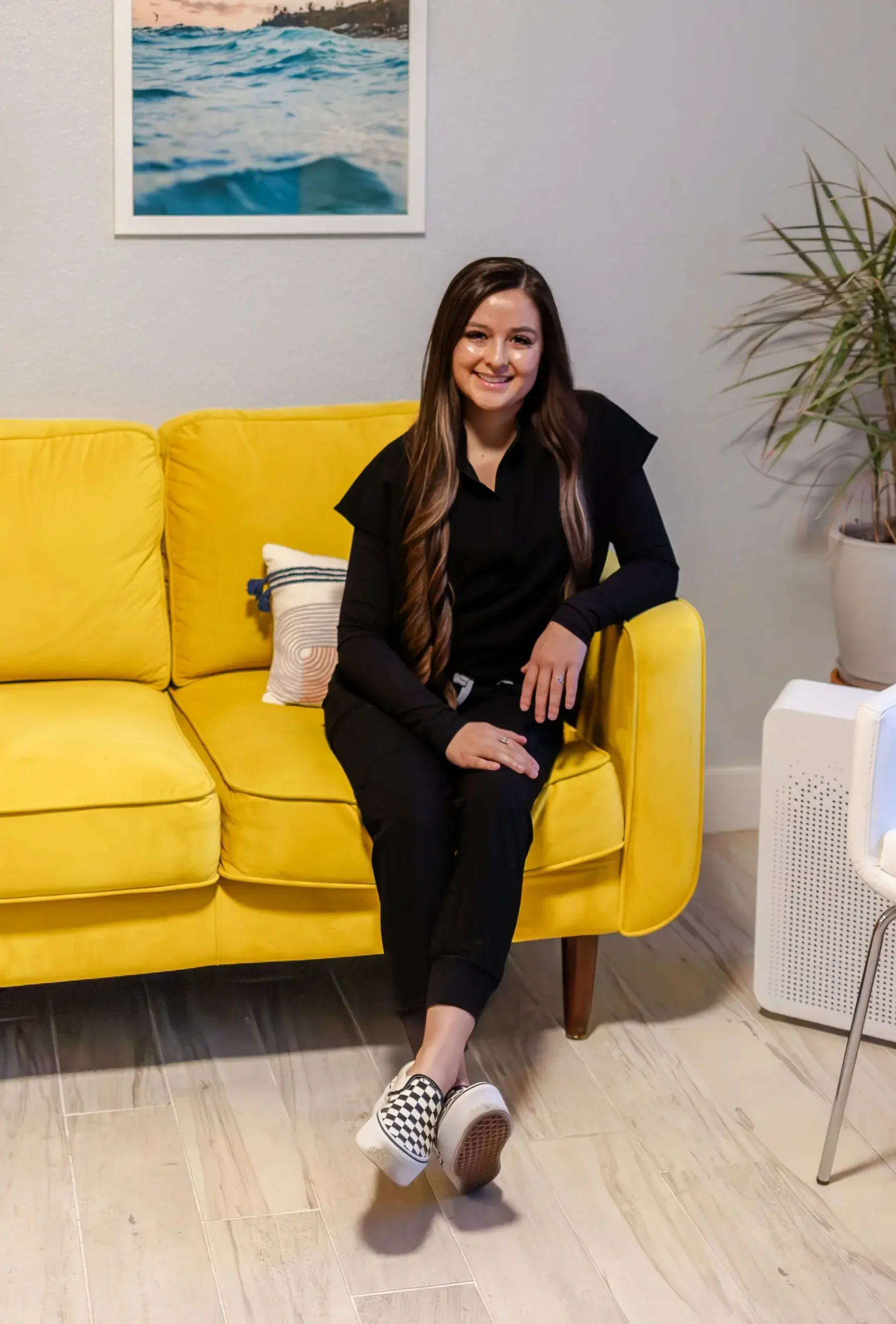 A woman with long, wavy brown hair, sitting on a bright yellow sofa in a living room, smiling at the camera, with a poster of water on the wall behind her and a potted plant on her right.