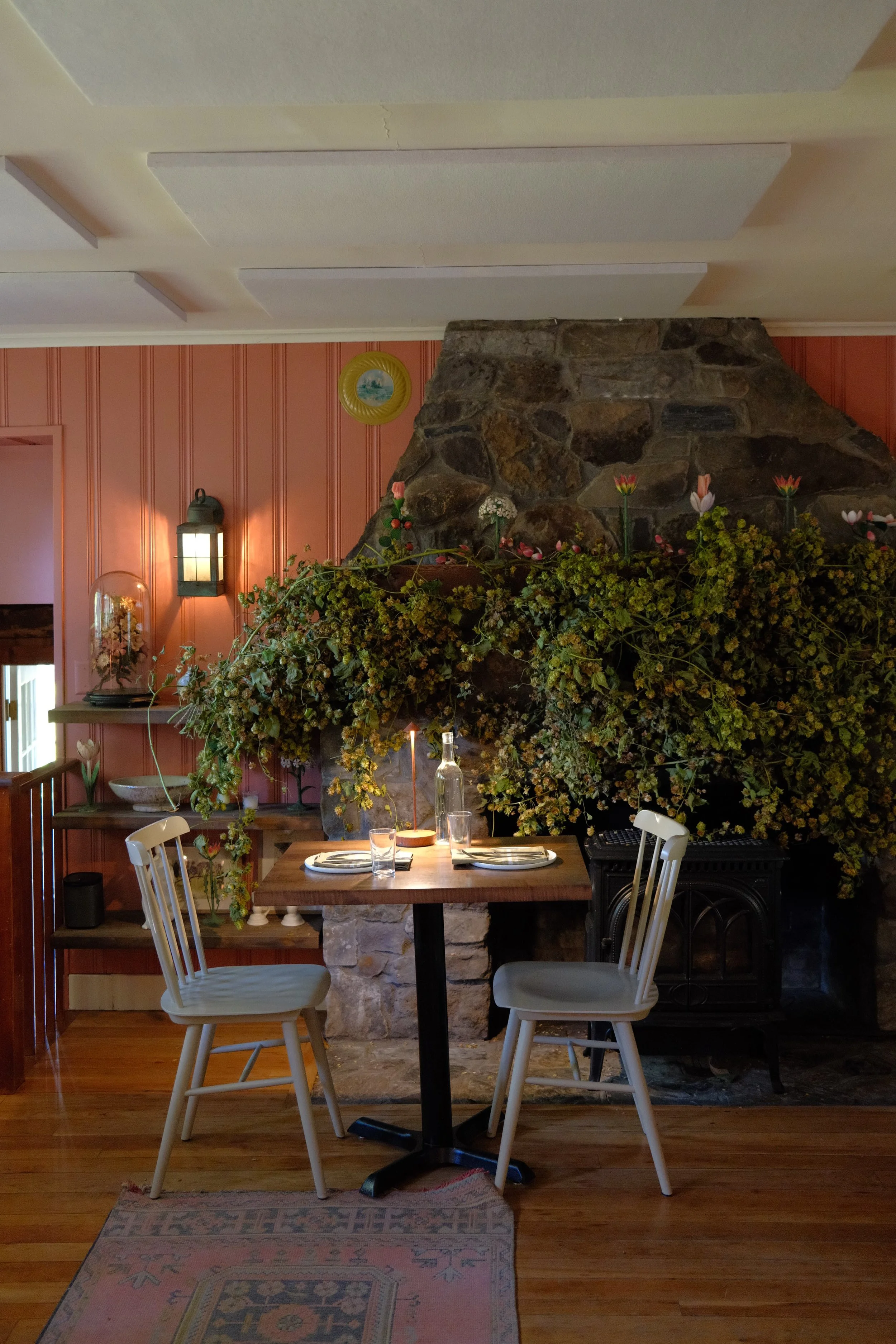 Indoor dining area with a wooden table set for two, two white chairs, a stone fireplace, decorative greenery, and a pink and orange striped wall with a yellow plate and wall-mounted lantern.