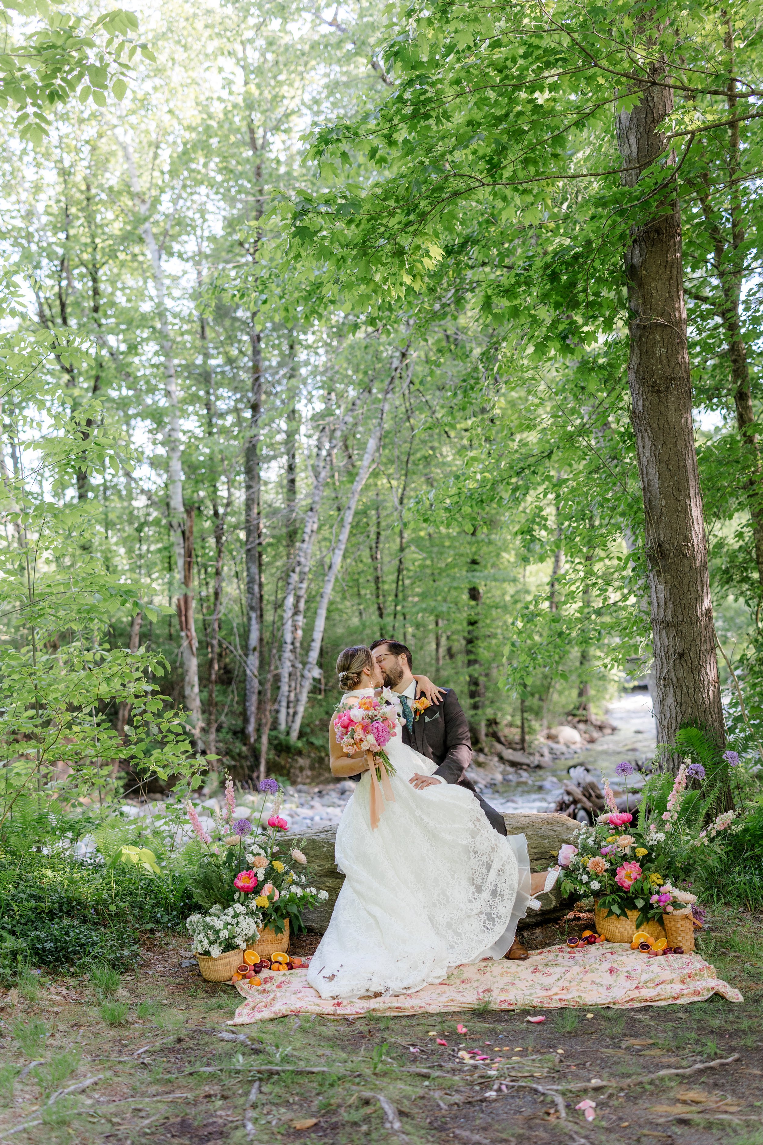 A newlywed couple sharing a kiss in a lush forest setting, surrounded by colorful flowers and baskets of fruit, on a picnic blanket by a small stream.