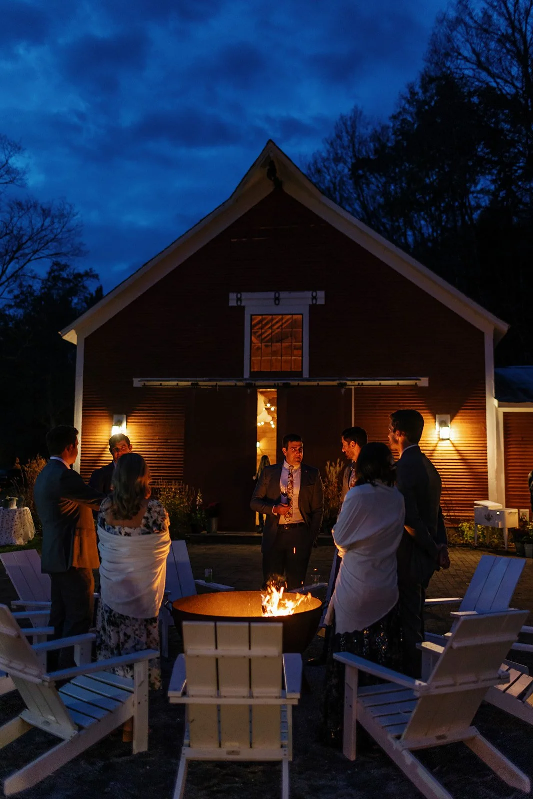 People gathered around a fire pit at night in front of a barn, with trees and a dark sky in the background at The Tillerman Inn in Bristol, VT.