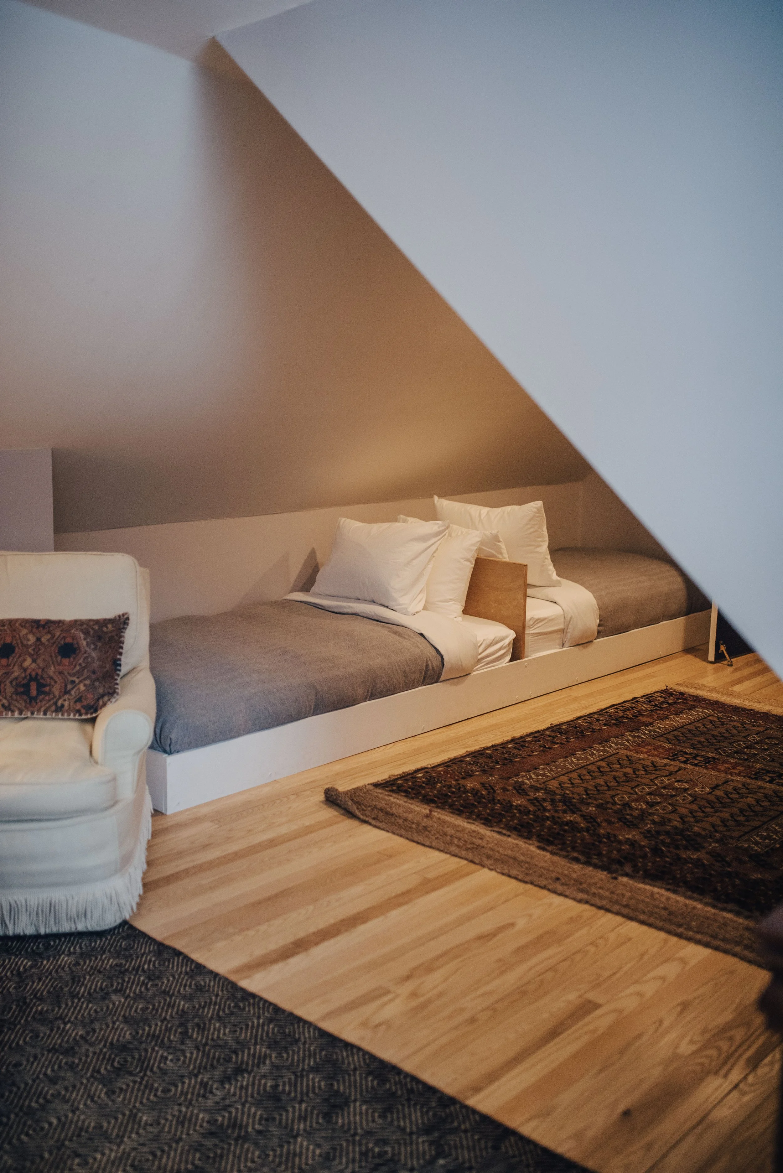 Cozy attic room with low bed against the wall, topped with white pillows and gray blanket. Armchair with patterned pillow next to bed, and traditional rug on the wooden floor at The Tillerman Inn in Bristol, VT.