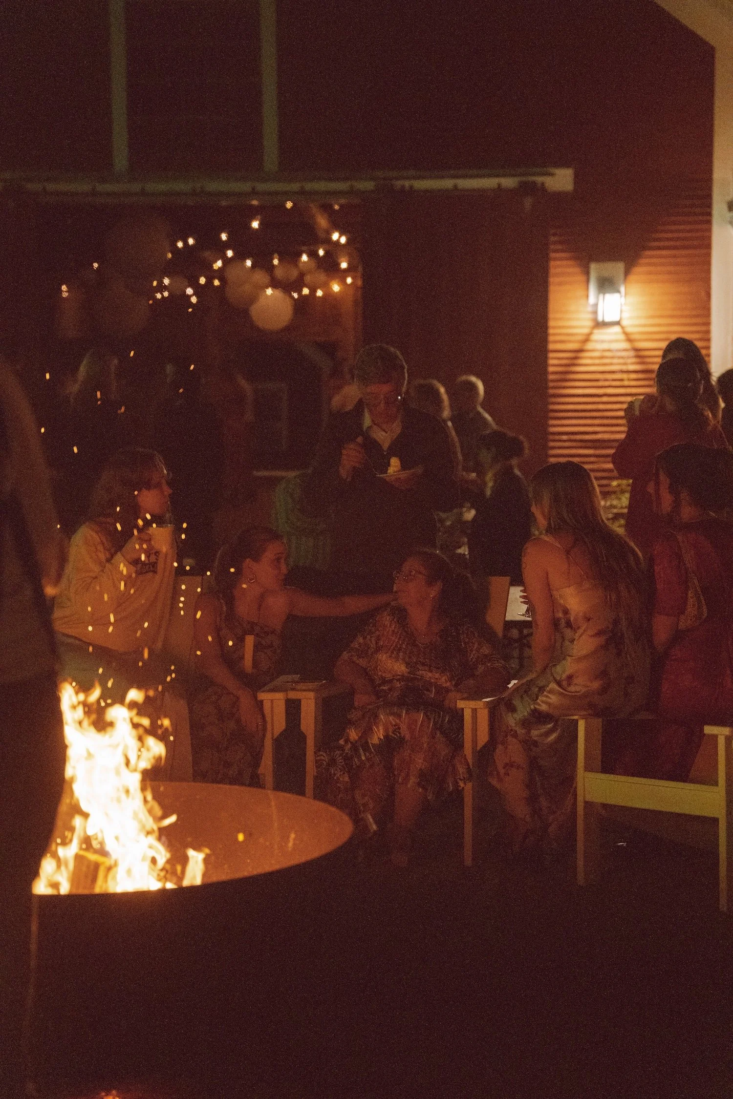 People gathered around a fire pit outside a rustic barn at night, with blue sky and trees in the background at The Tillerman Inn in Bristol, VT.
