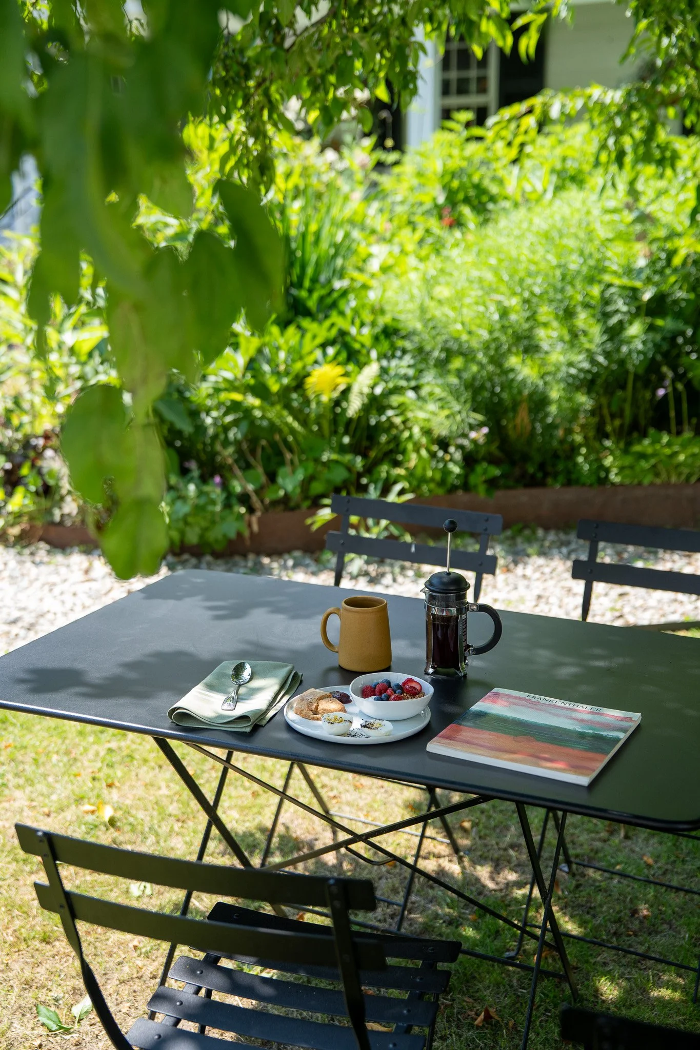 Outdoor table set for breakfast with a plate of berries, croissant, and yogurt, a cup, a French press, a book, and utensils on a green table in a garden with lush green bushes and trees in the background.