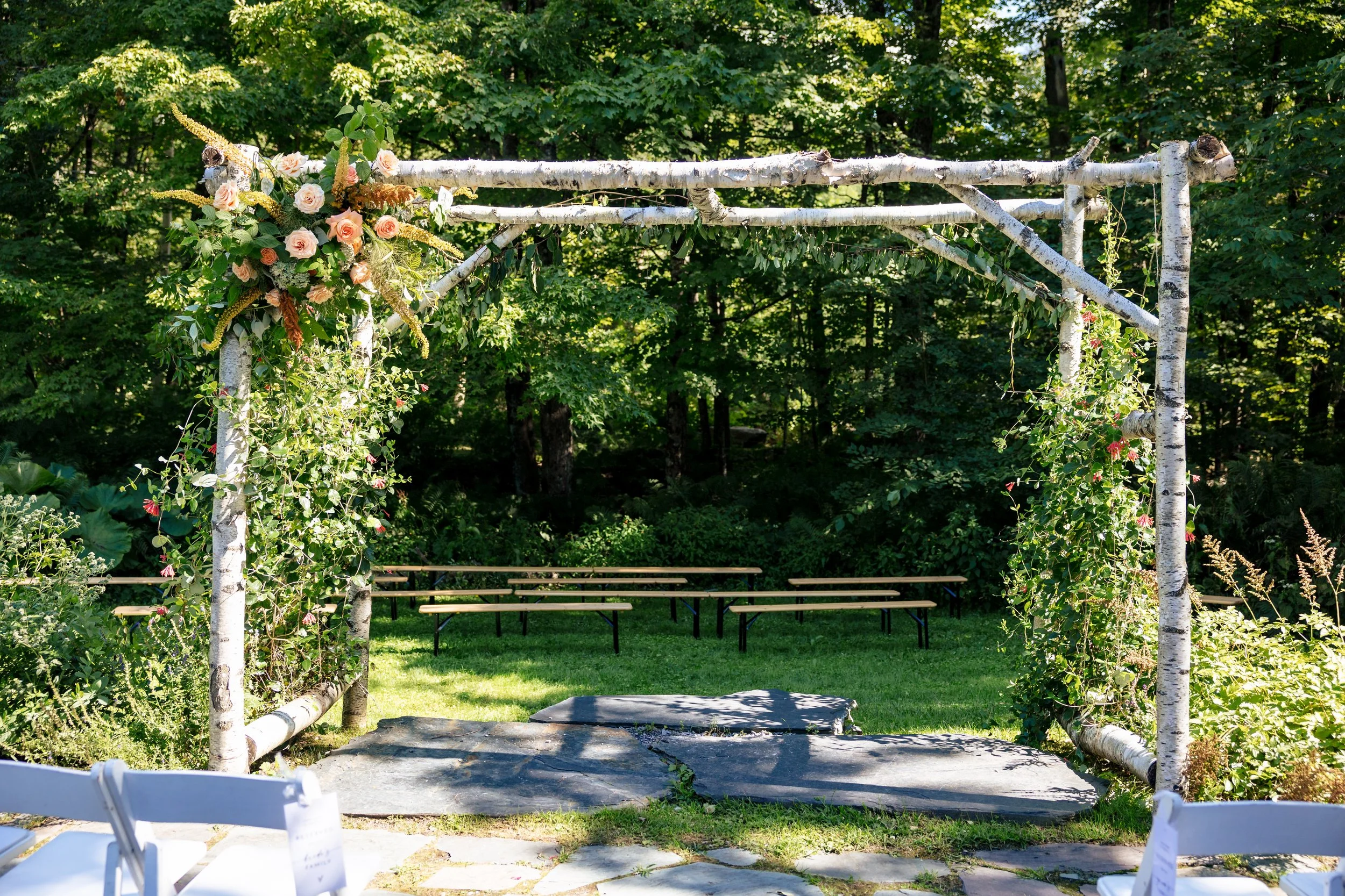 a birch arch adorned with flower arrangements at our ceremony site on the stone patio overlooking the forest at The Tillerman in Bristol, Vermont.