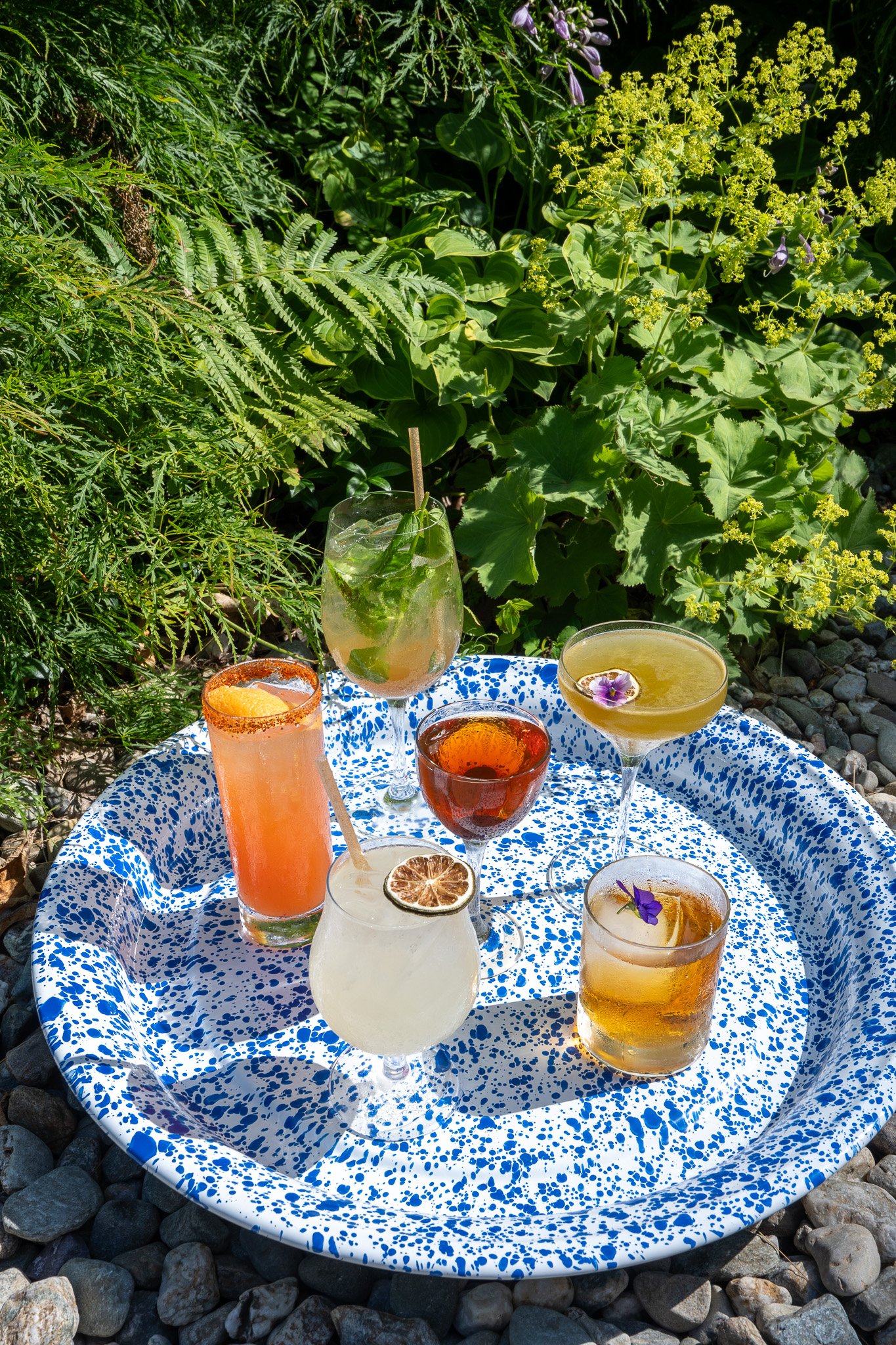 speckled blue and white enamel tray carrying 6 colorful summery cocktails sitting beside the garden in the patio at The Tillerman in Vermont