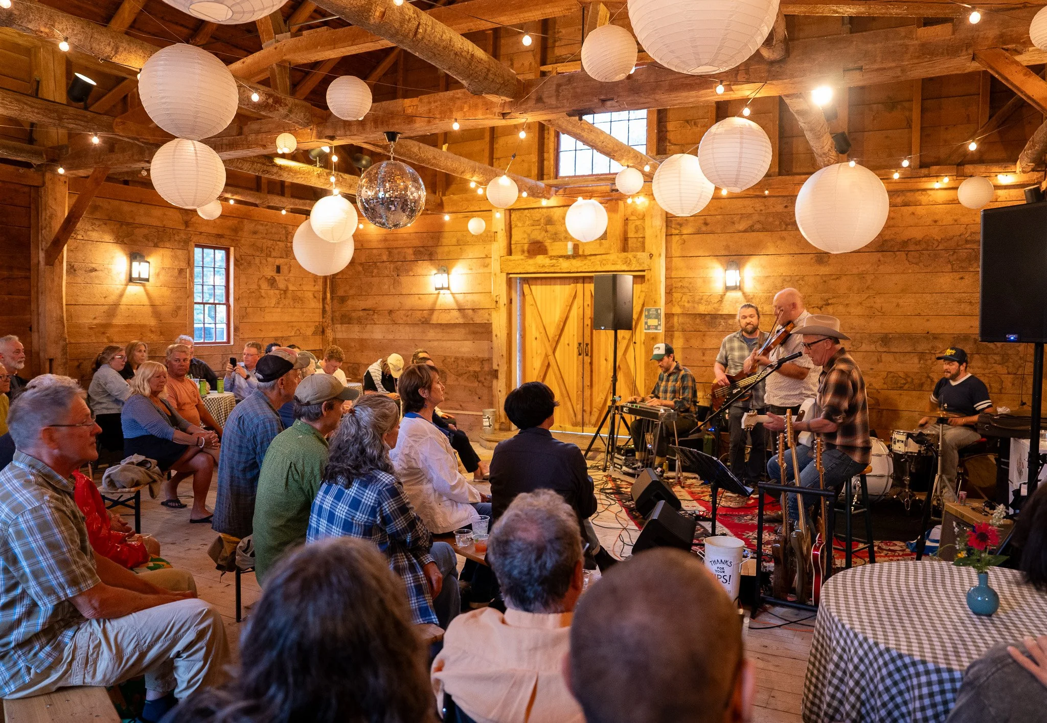 a bluegrass plays for a large crowd in the historic vermont barn with paper lanterns and twinkling hanging from the ceiling