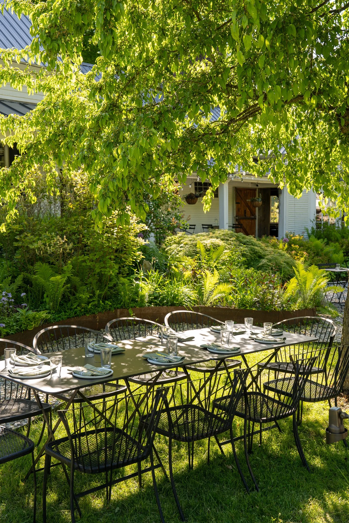 the outside of the tillerman, an old white vermont farmhouse, in summertime on the patio by the gardens. dappled light on the large outdoor table.