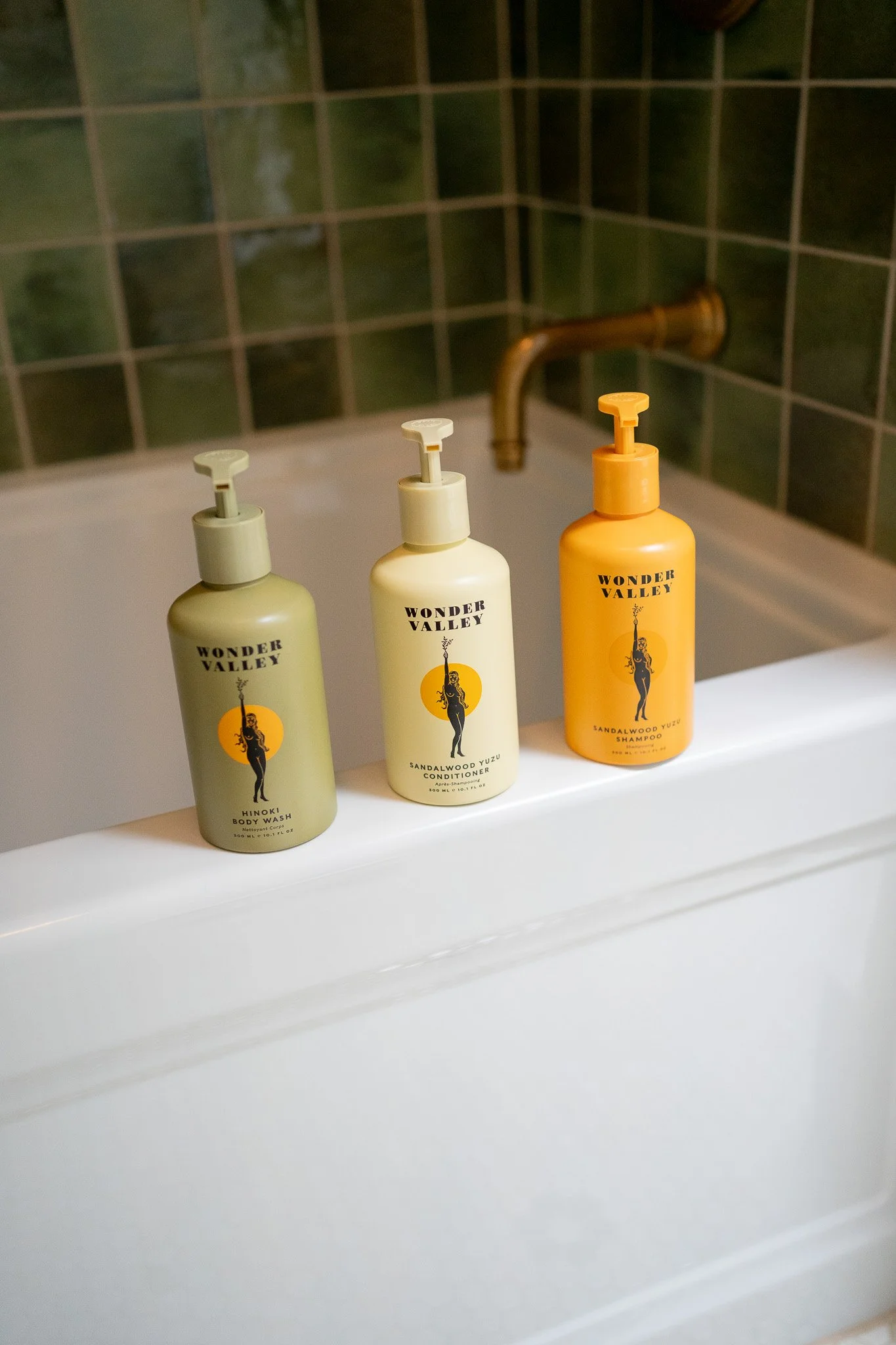 wonder valley hair and body care products resting on the side of the en-suite tub in North Country. A brass bath faucet and green tile is behind