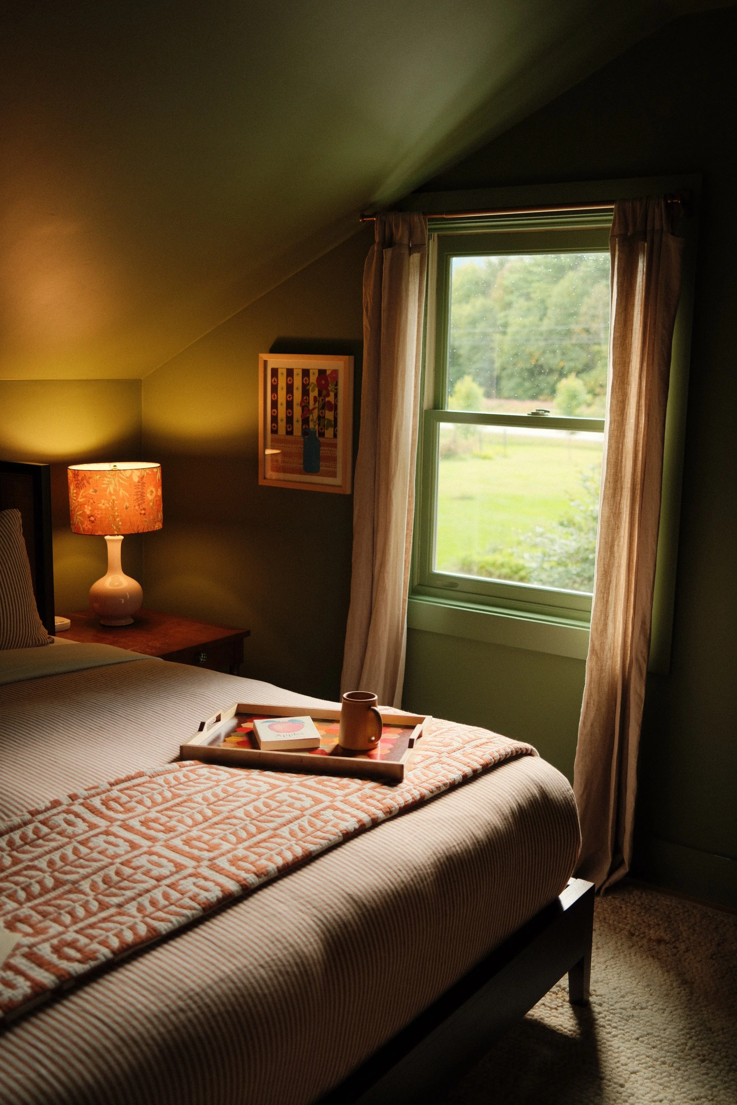 A bedroom with a bed, nightstand, table lamp, window with curtains showing greenery outside, and a tray with a mug and a book on the bed.