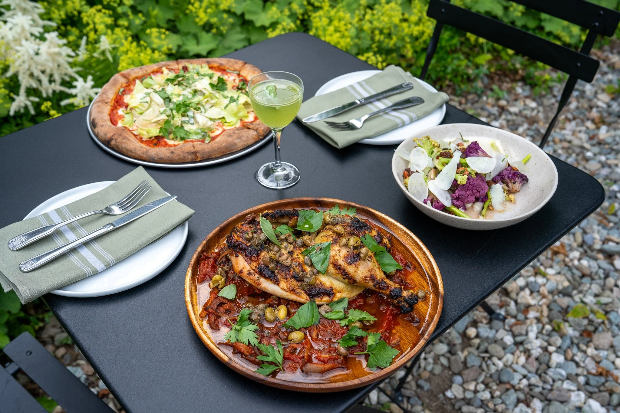 the outside of the tillerman, an old white vermont farmhouse, in summertime on the patio by the gardens. shared plates sit on the table with a festive cocktail. 