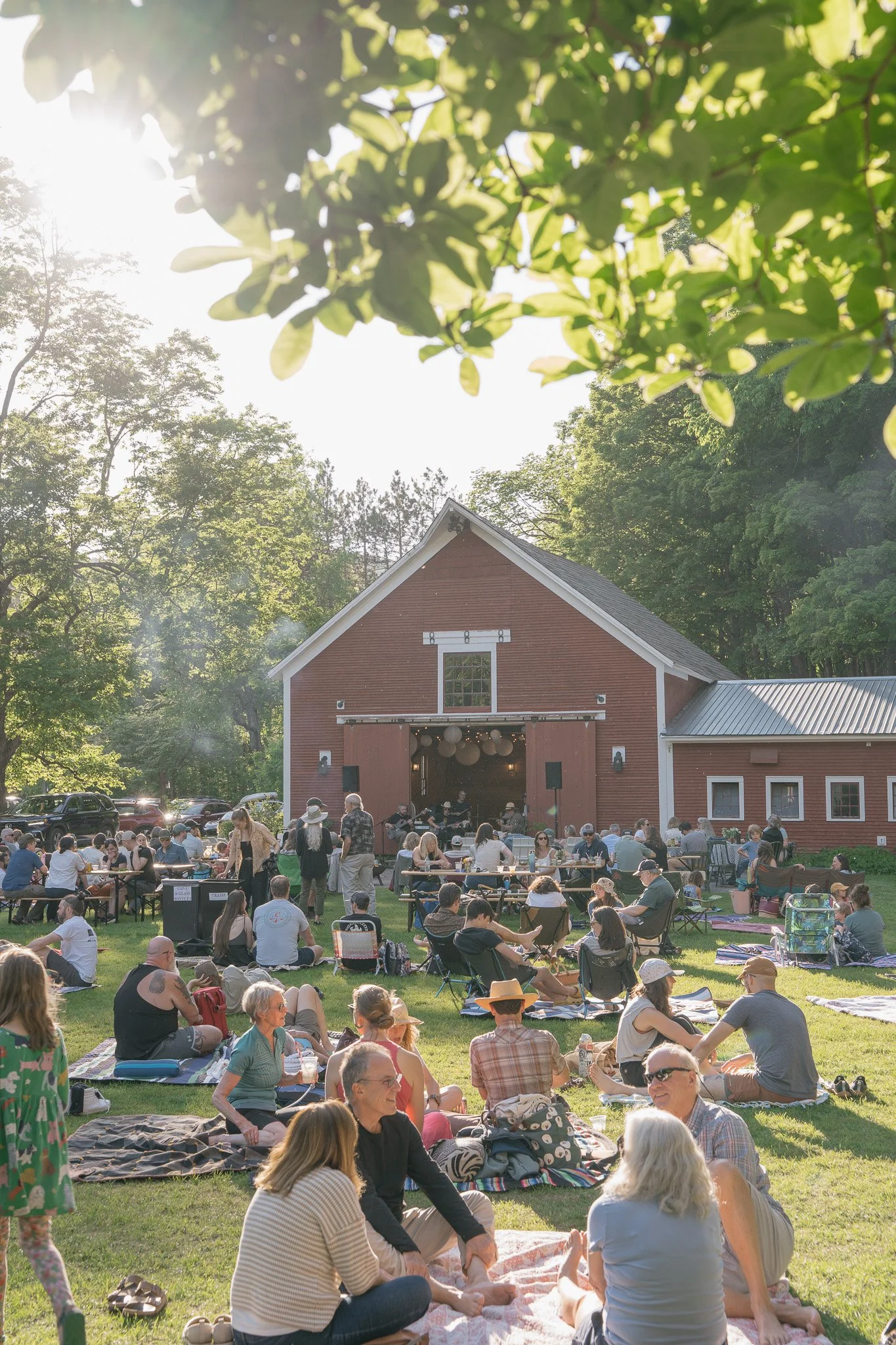 People gathered outside the red barn at The Tillerman Inn in Bristol, VT for an outdoor event with a stage, some sitting on blankets and chairs on the grassy area and others standing or walking. The event is in a sunny, green park.