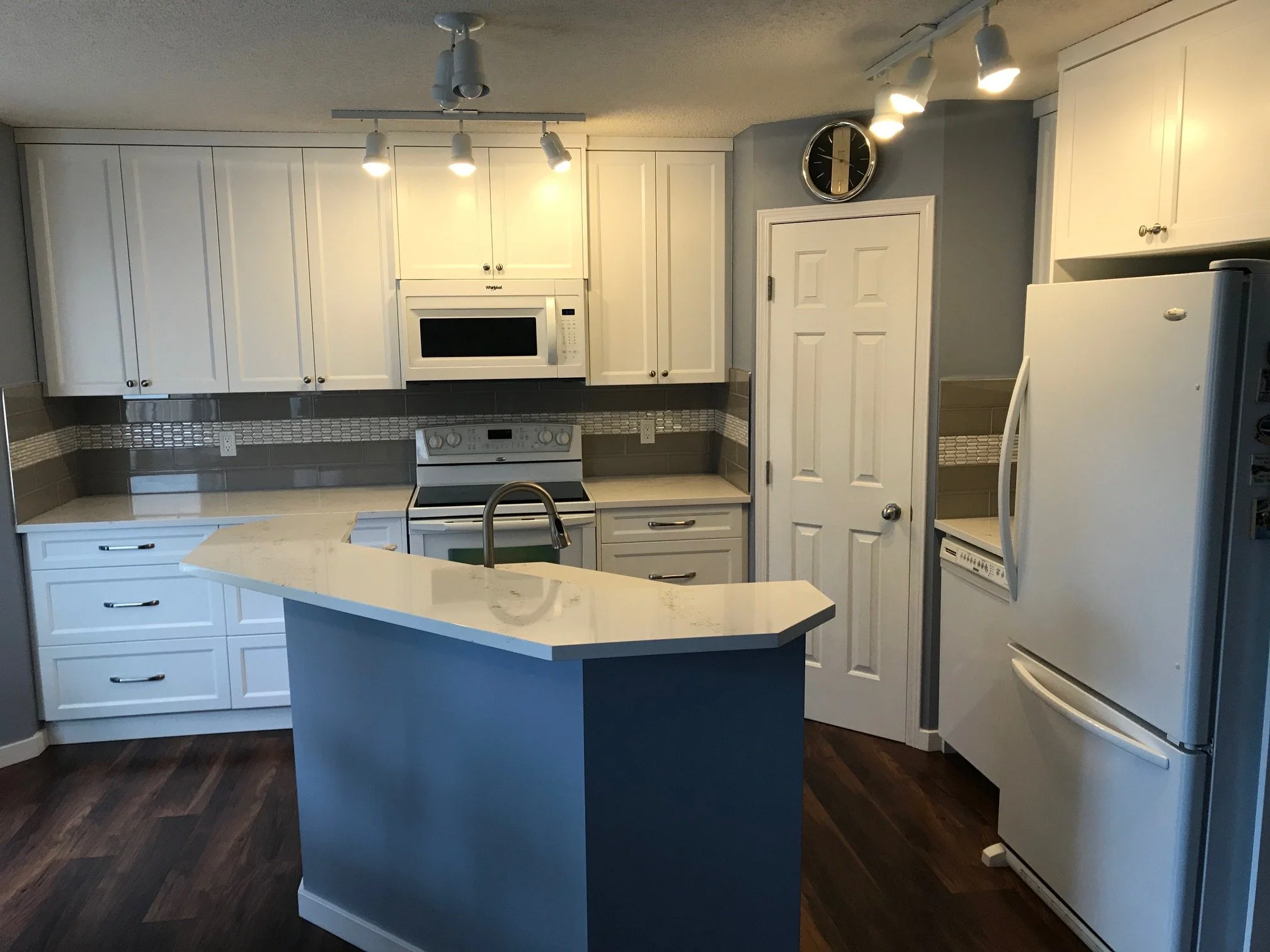 Clean modern kitchen with white cabinets, a microwave, stove, refrigerator, small island with a sink, and a wall-mounted clock.