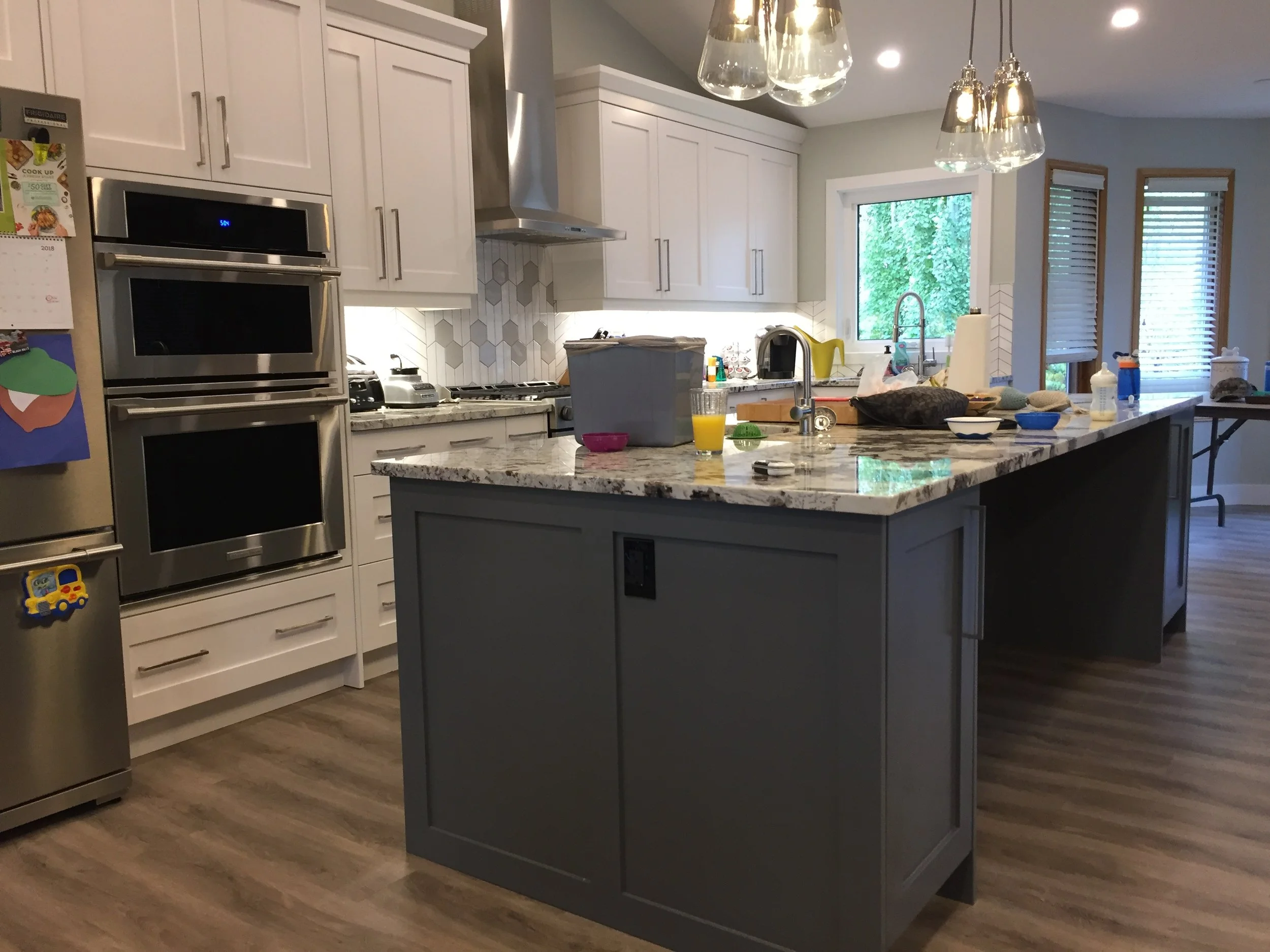 Modern kitchen with white cabinets, a gray island, stainless steel double oven, and a marble countertop. There are windows with blinds and hanging pendant lights.