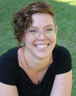 A smiling woman with glasses and short hair, outdoors on grassy ground.