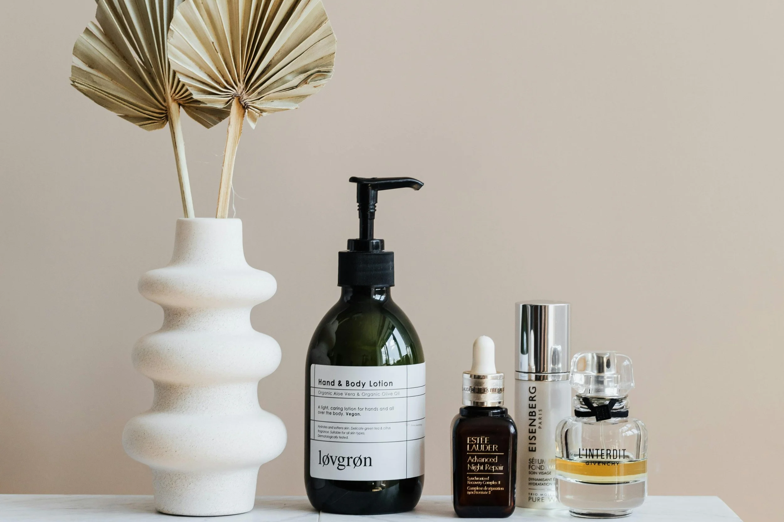 Hintergrund beige, weiße Tischfläche, weiße Vase mit getrockneten Palmblättern, Dunkle Flasche Hand- und Körperlotion, kleinere Flasche mit Pipette, silberne und gläserne Parfümflaschen.