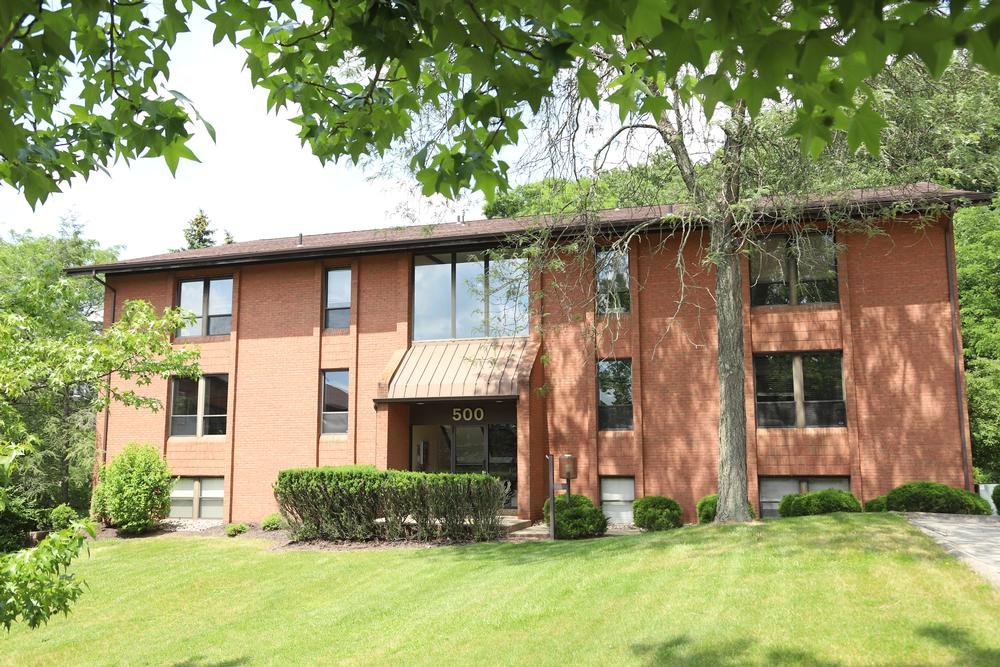 Two-story brick apartment building with green lawn and trees, address number 500 above the entrance, and large windows reflecting the sky.