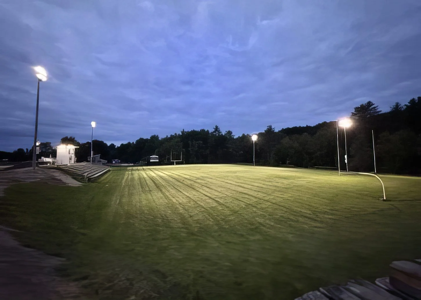 A baseball or softball field during evening or early night with bright floodlights illuminating the field, surrounded by trees and a partly cloudy sky.