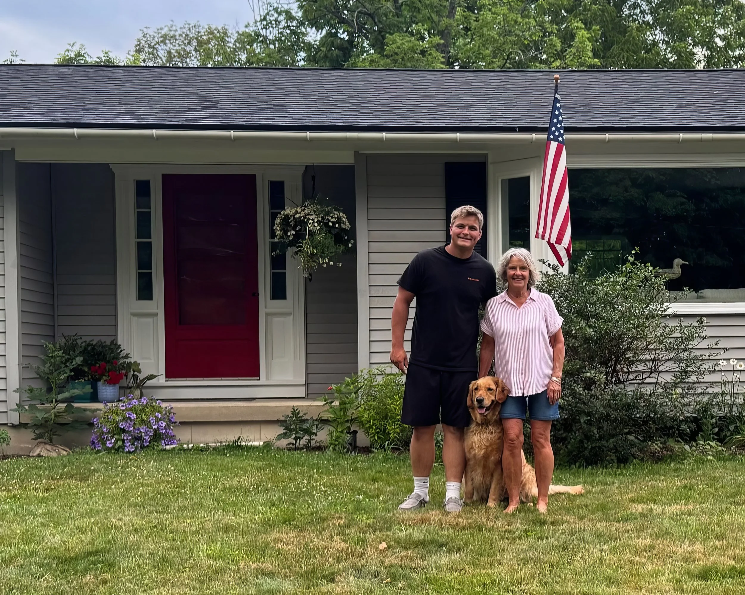 A man, woman, and golden retriever standing in front of a house with a red door, American flag, and garden flowers.