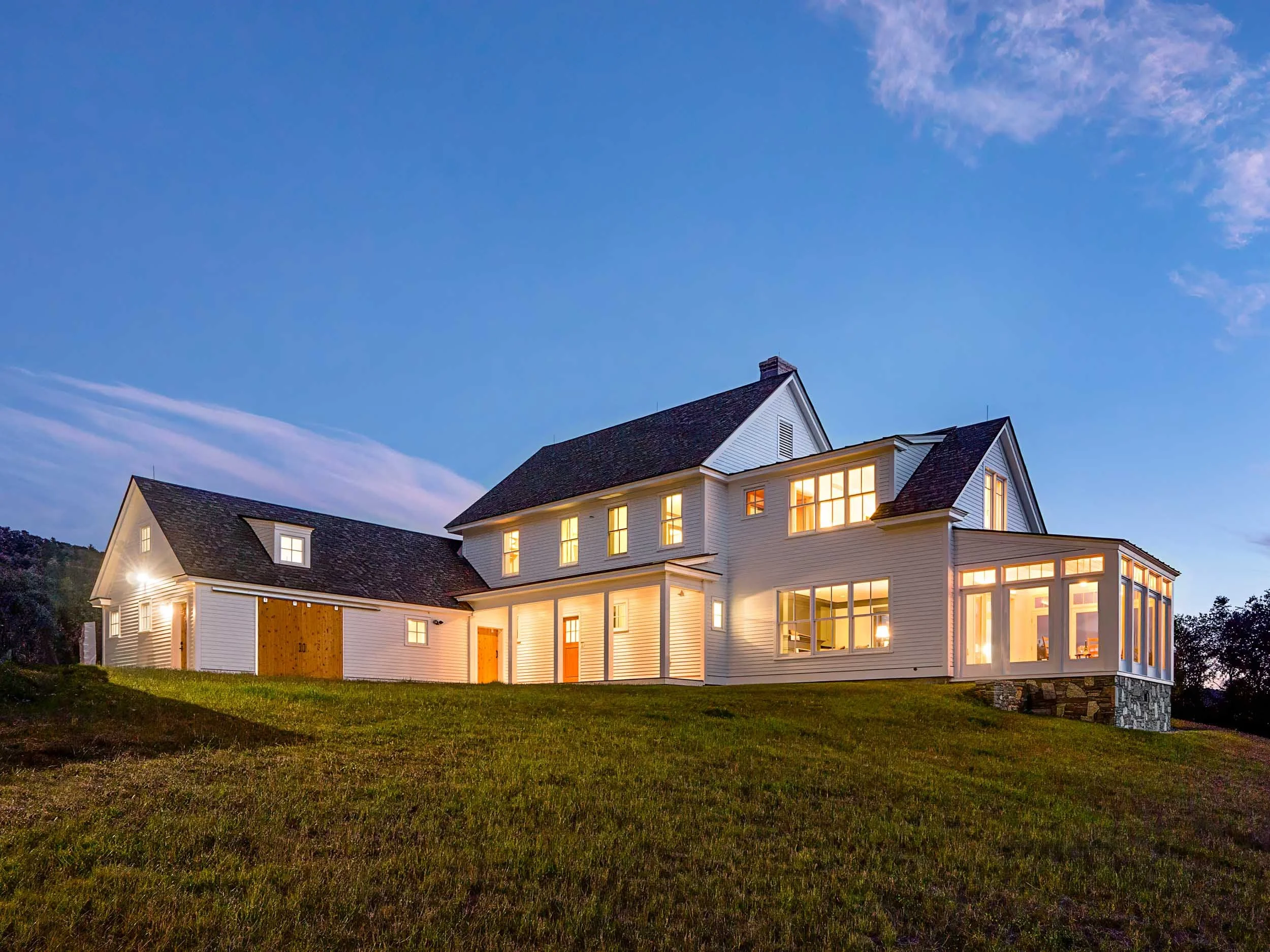 A large white house with multiple levels and large windows, illuminated from inside, on a grassy hill at dusk.