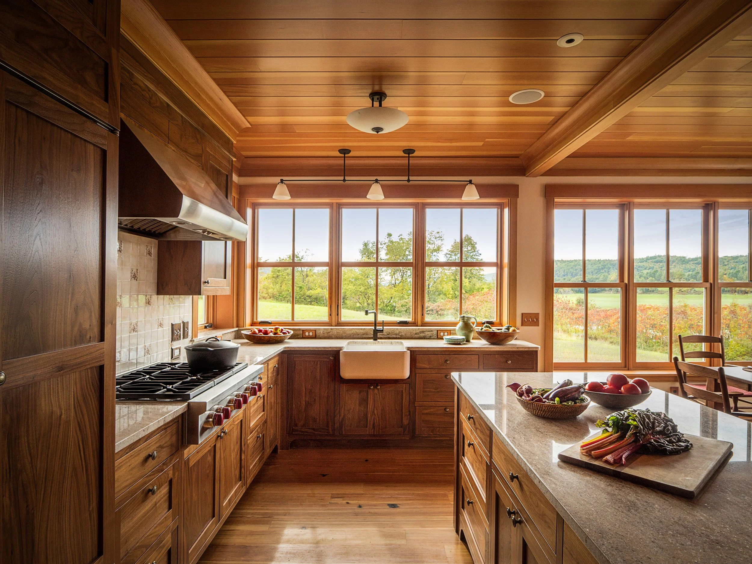 A bright kitchen with wooden cabinets and large windows overlooking a green landscape, with a countertop featuring a bowl of fruit, a cutting board with vegetables, and a dining table with chairs.