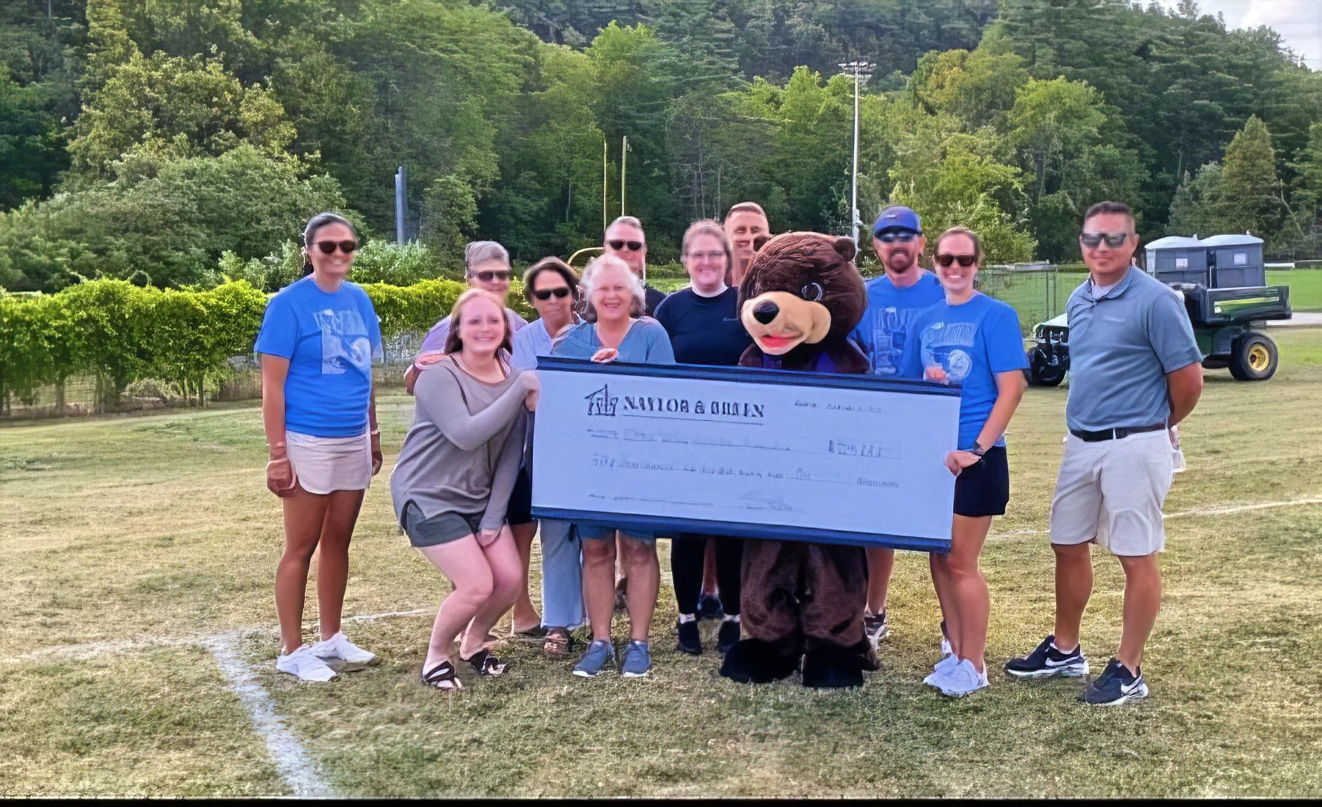 Group of people on a football field holding a large check, with a person in a bear mascot costume in the center, and trees in the background.
