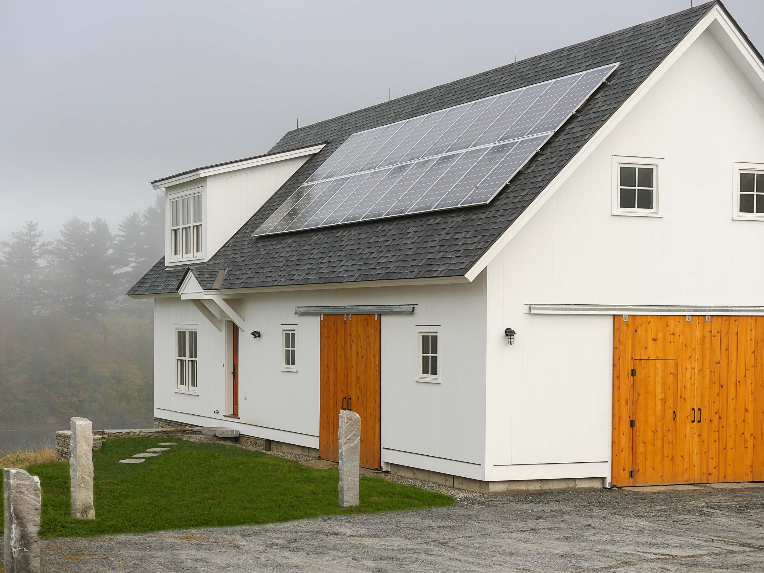 A white two-story house with a black shingled roof, equipped with solar panels, a wooden garage door, and several windows, situated in a foggy outdoor setting with a gravel driveway and grassy area.