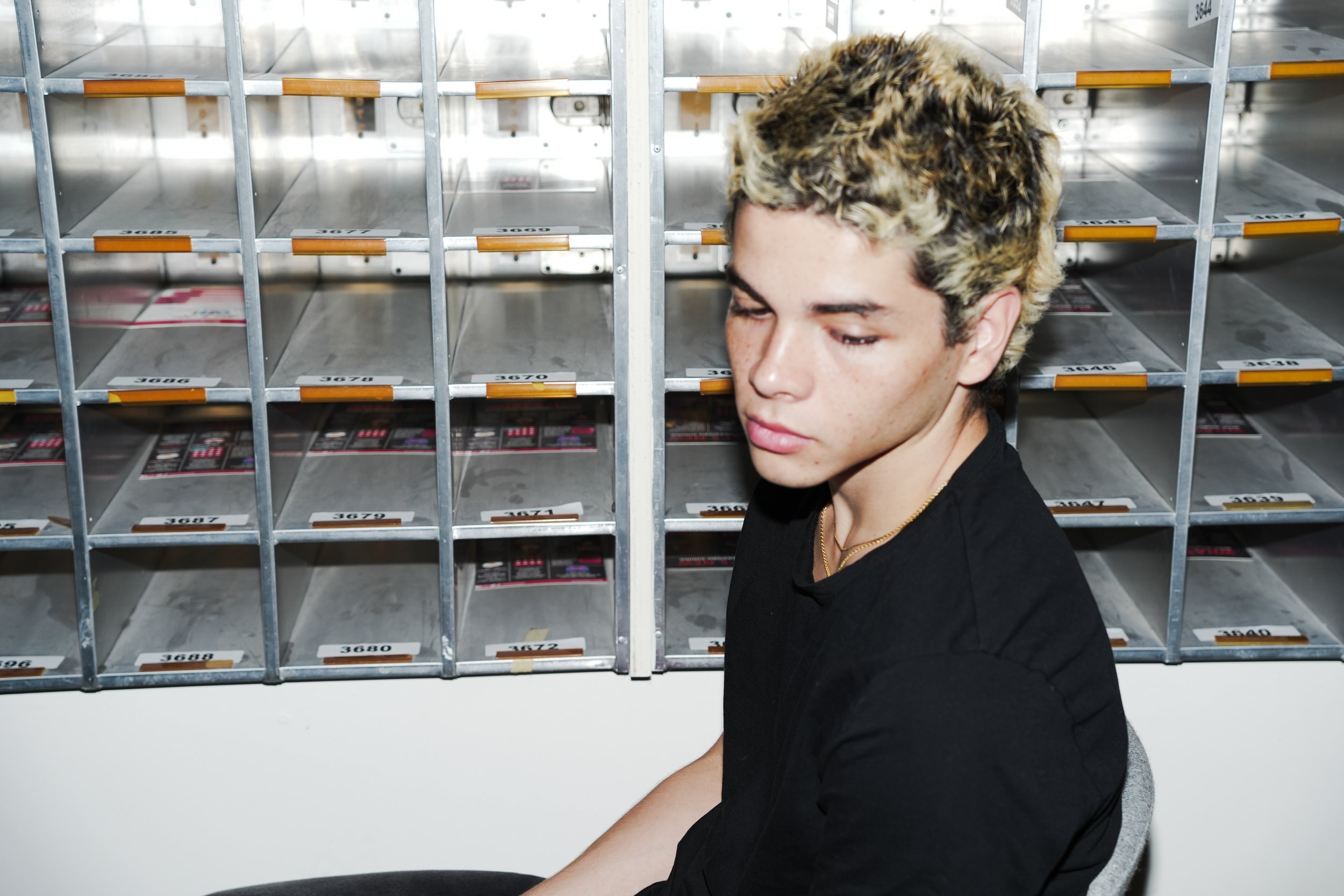 Young man with blond hair sitting in front of empty mailboxes or storage cubbies.