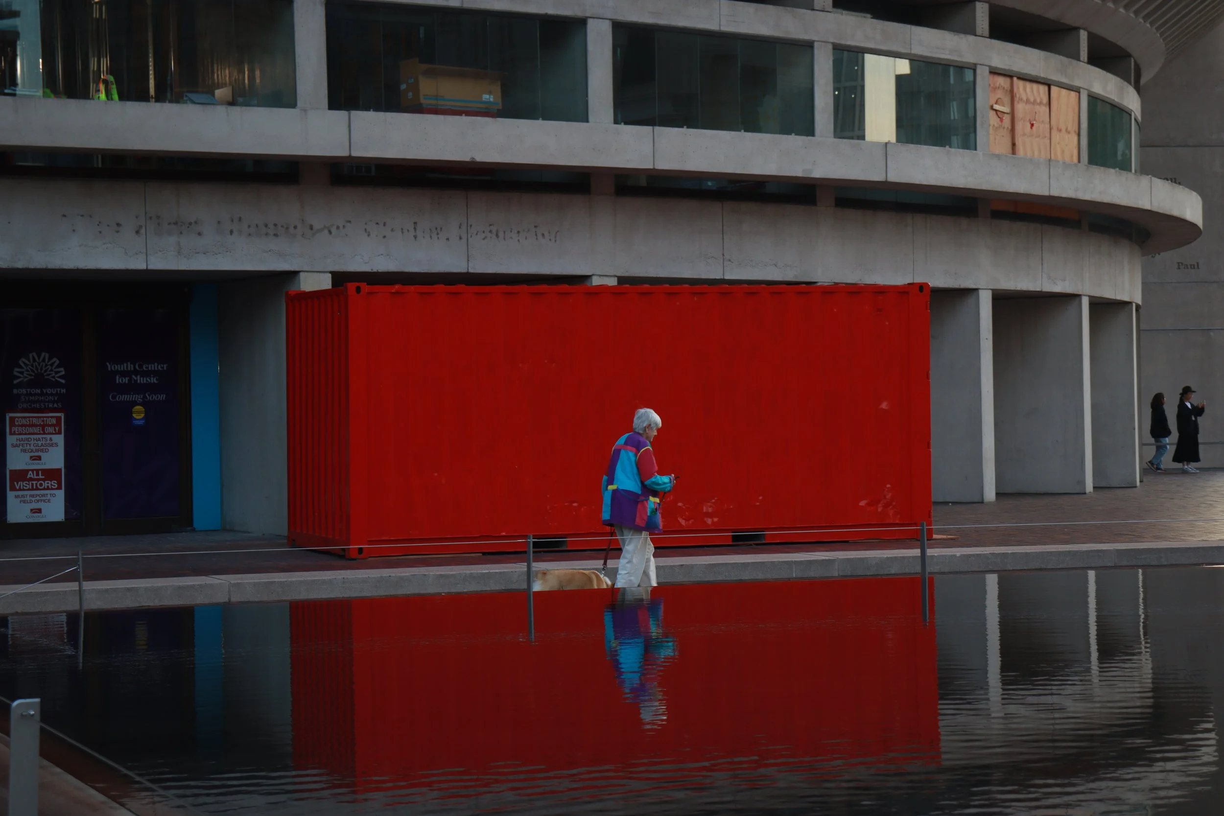 An elderly woman in a colorful jacket walking her dog along the edge of a reflecting pool in an urban area, with a modern building behind her and two other people taking photos in the background.