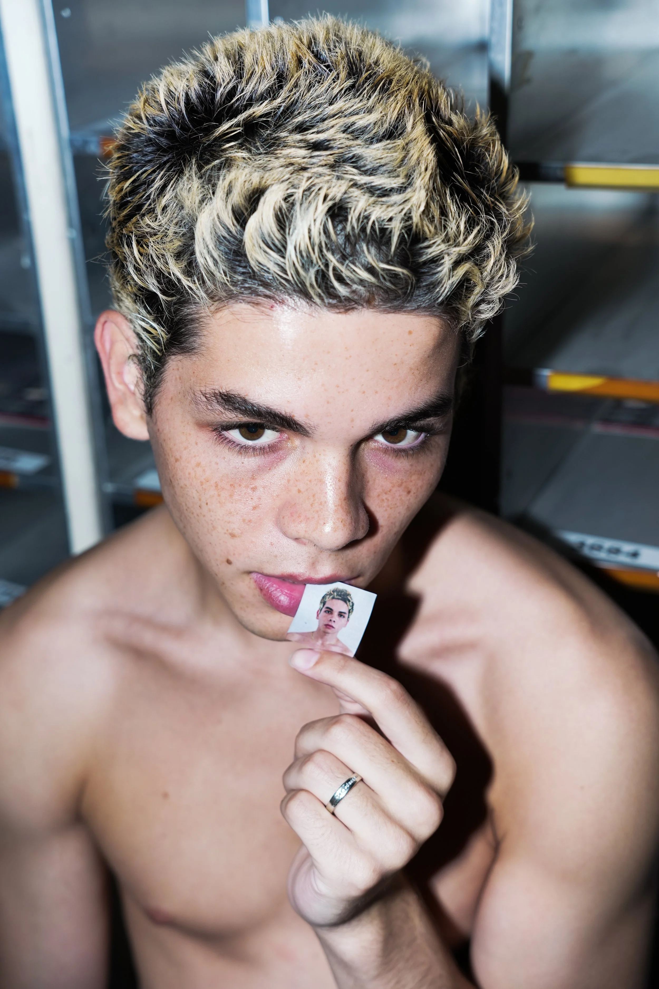 A shirtless person with short, curly blond and dark hair holding a small photo of themselves near their lips, looking directly at the camera.