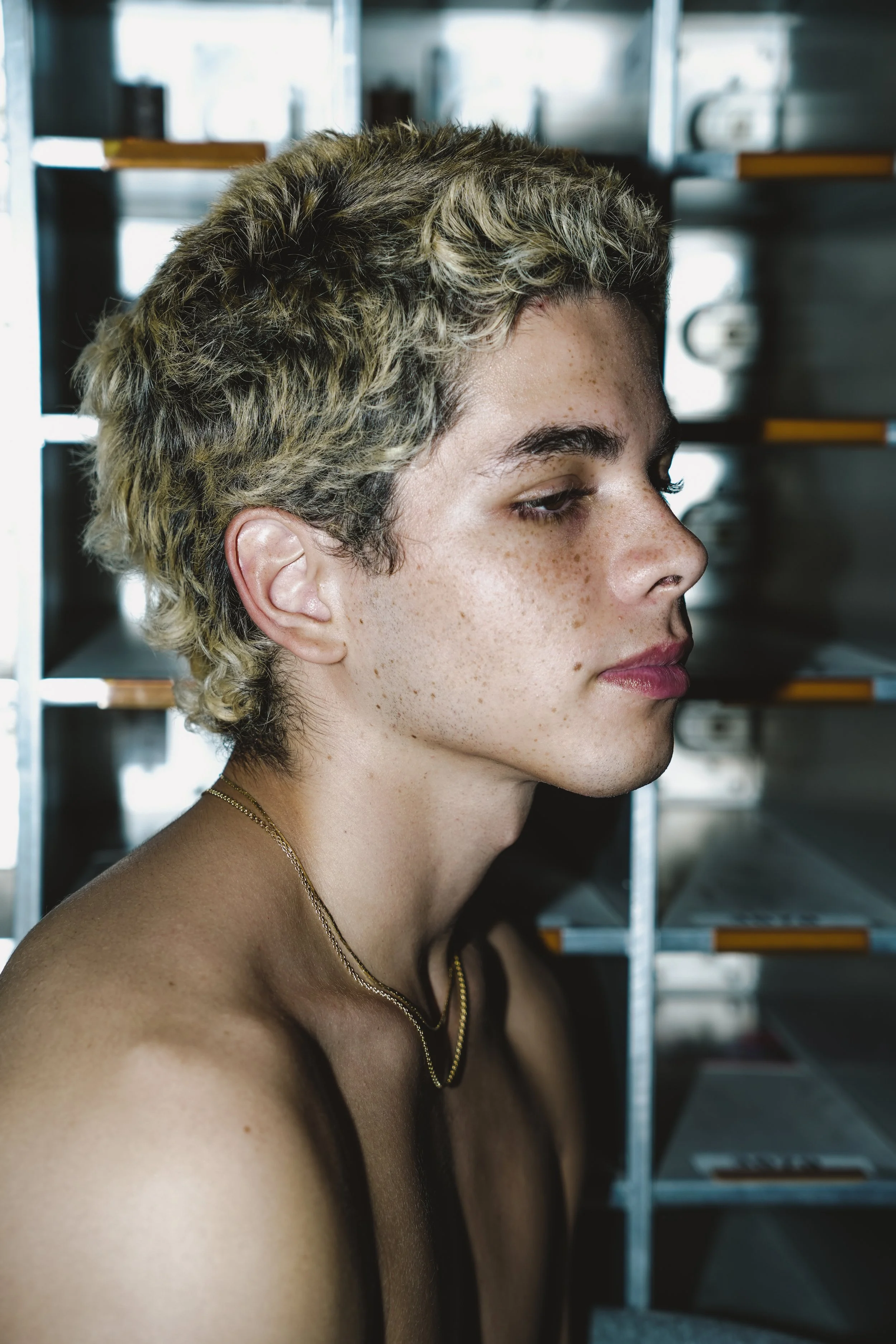 Profile of a person with short, curly blonde hair, wearing a gold chain necklace, standing in front of metal shelves with trays or boxes.
