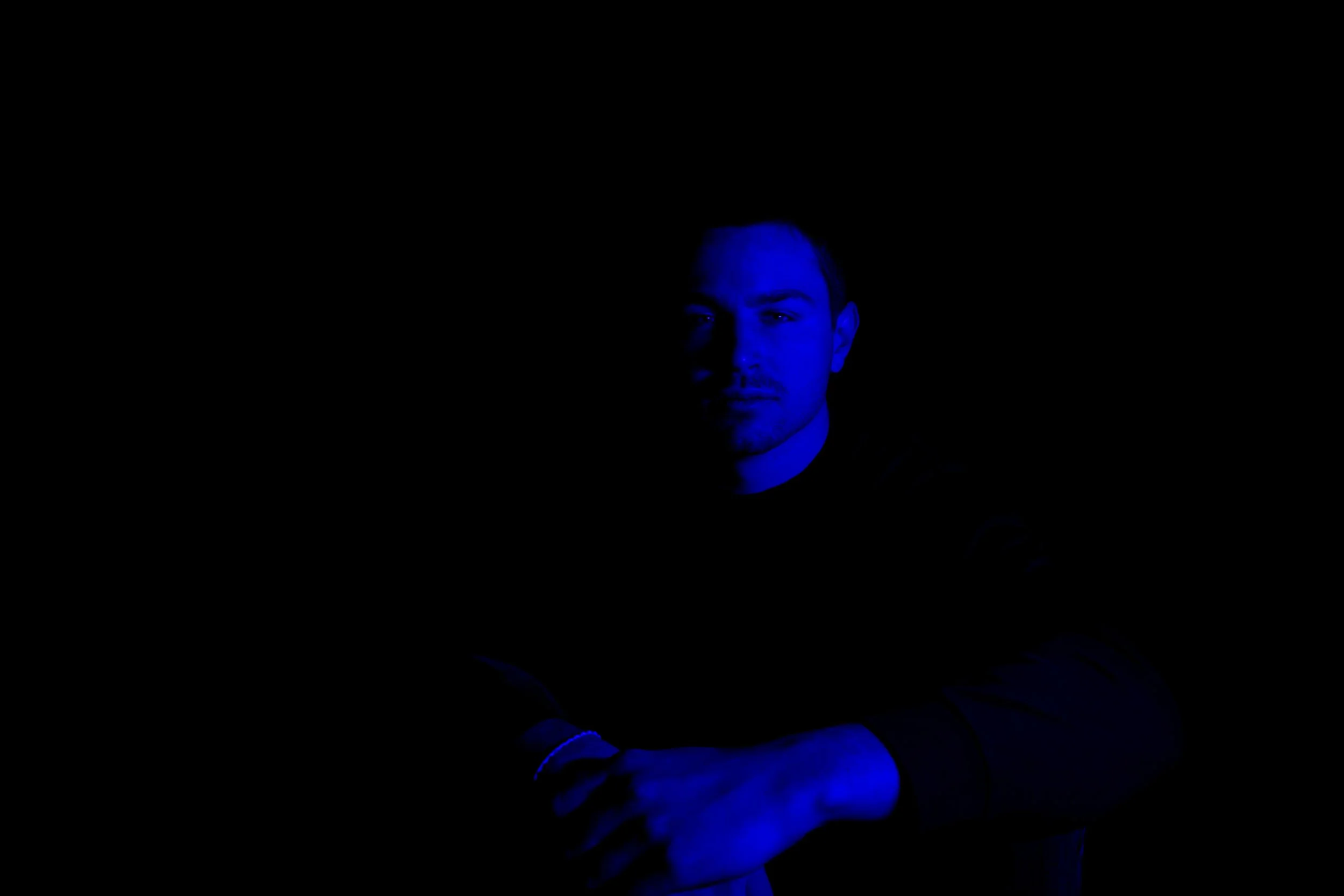A portrait of a man illuminated by blue lighting against a black background, with serious facial expression and arms crossed.