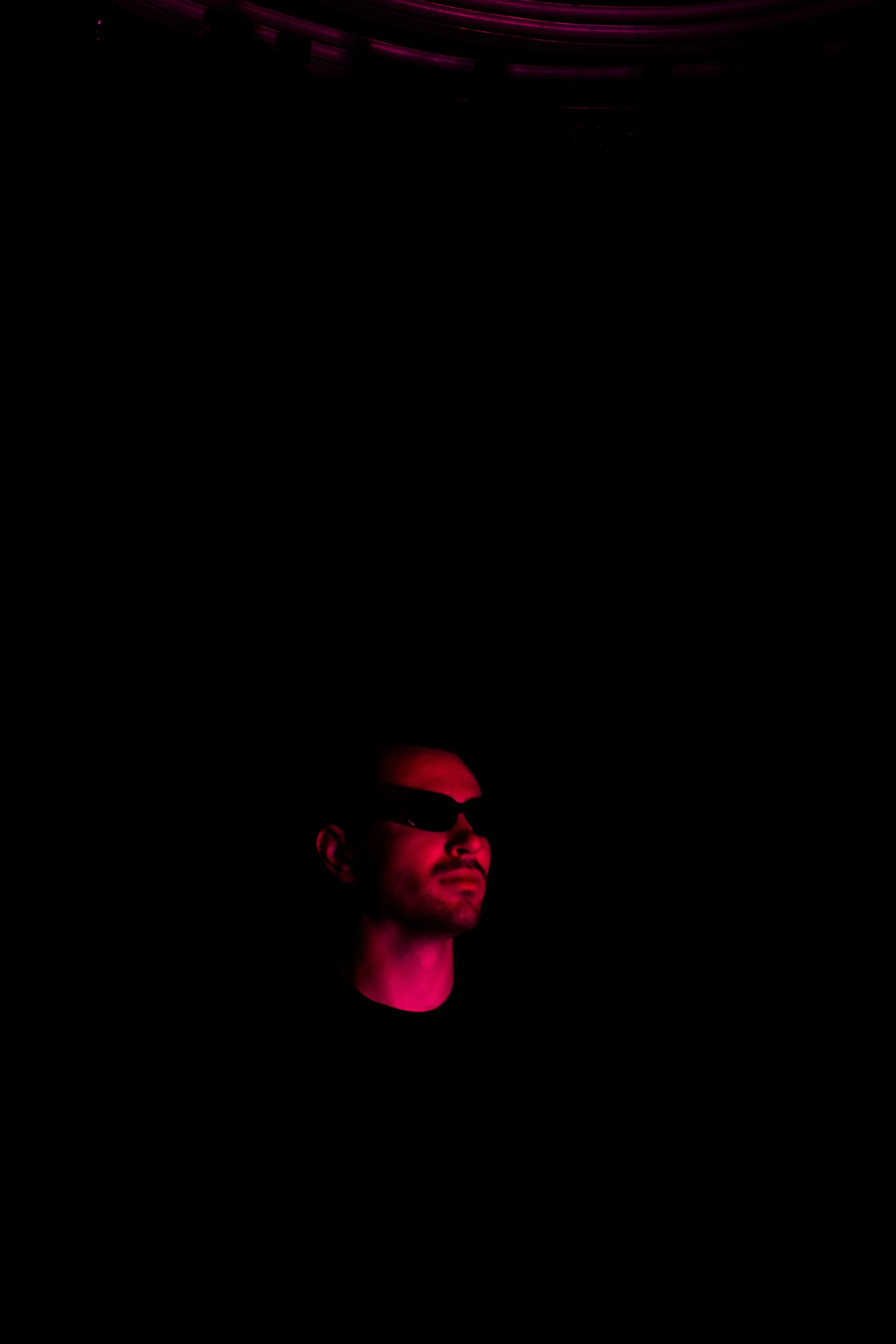 Portrait of a man wearing sunglasses, illuminated with pink and purple lighting, against a dark background.