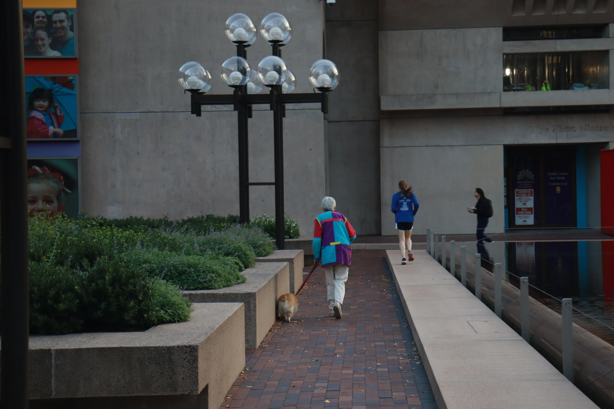 A person walking a dog along a paved path next to a modern building and a water feature, with another person jogging and a woman standing nearby, during daytime.