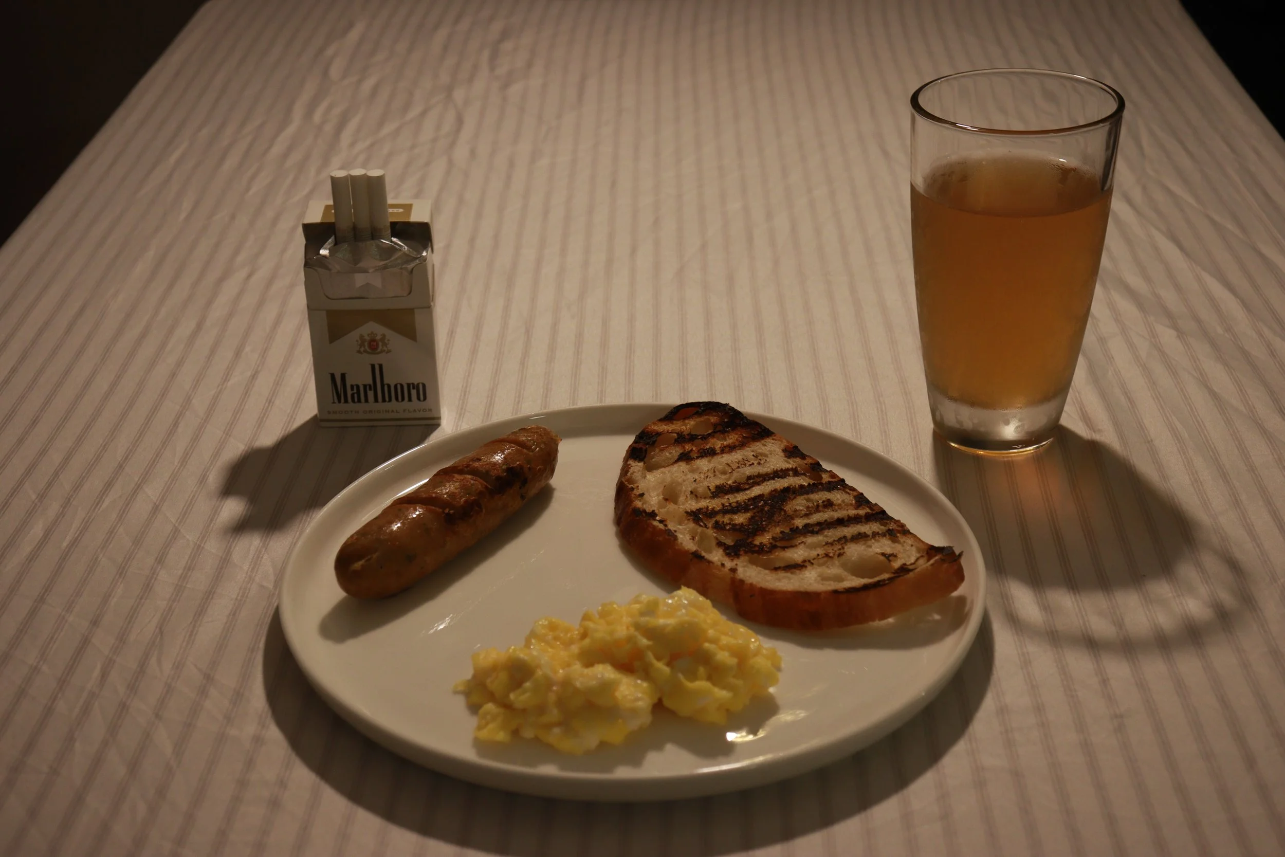 A breakfast plate with scrambled eggs, a grilled slice of bread, and a sausage link, along with a glass of orange juice, a pack of Marlboro cigarettes, on a white tablecloth.