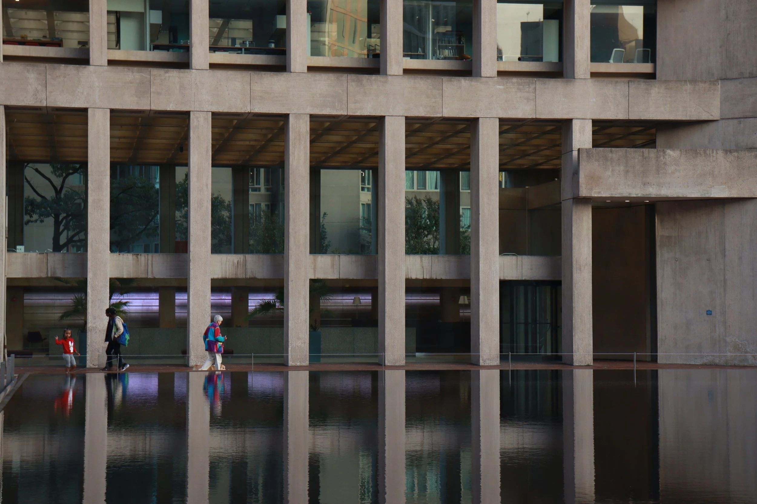 Three people walking along a reflective pool in front of a modern building with large glass windows and concrete columns.