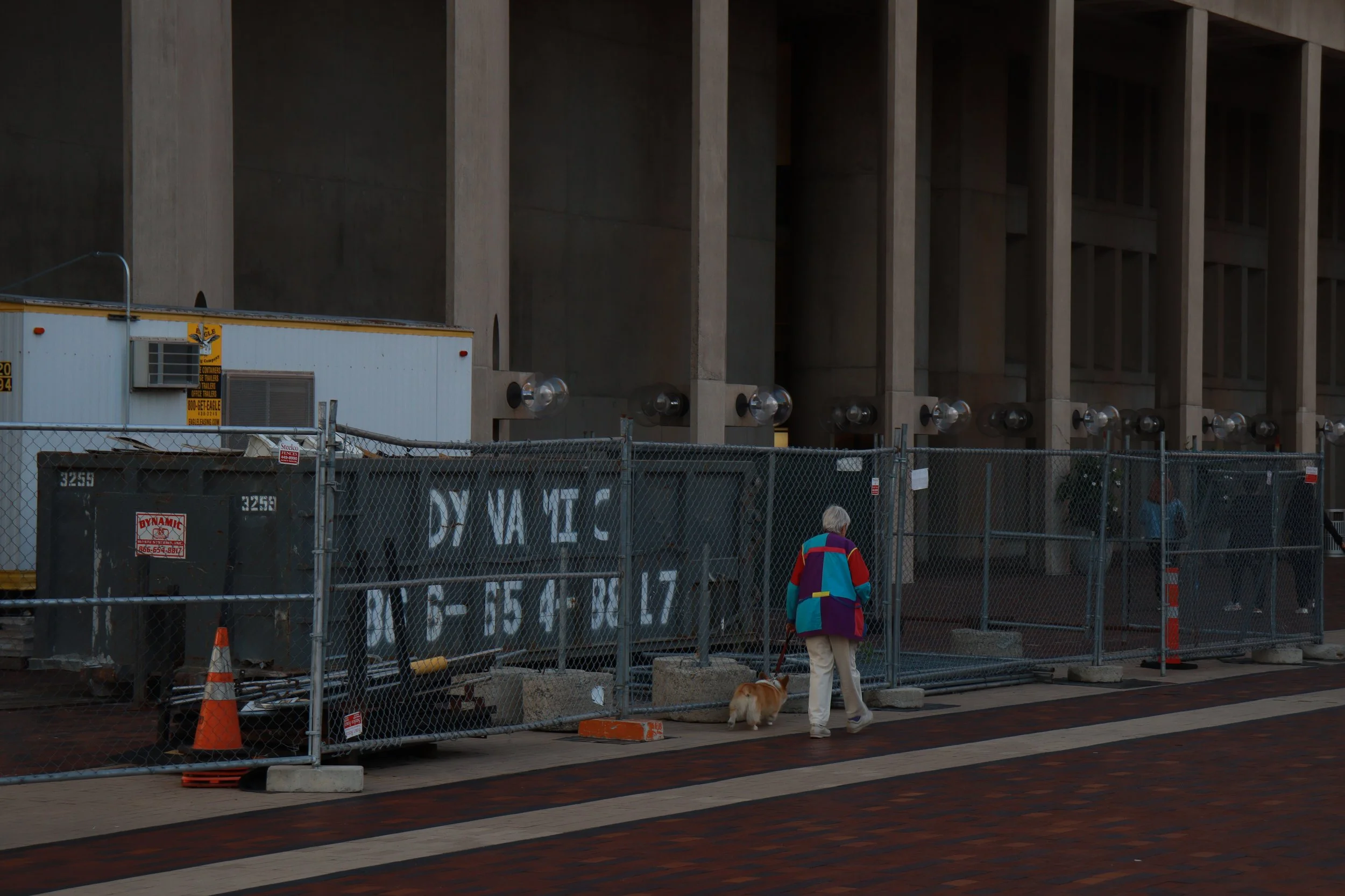 A person walking a dog past a construction site with chain-link fencing, construction equipment, and a large building in the background.