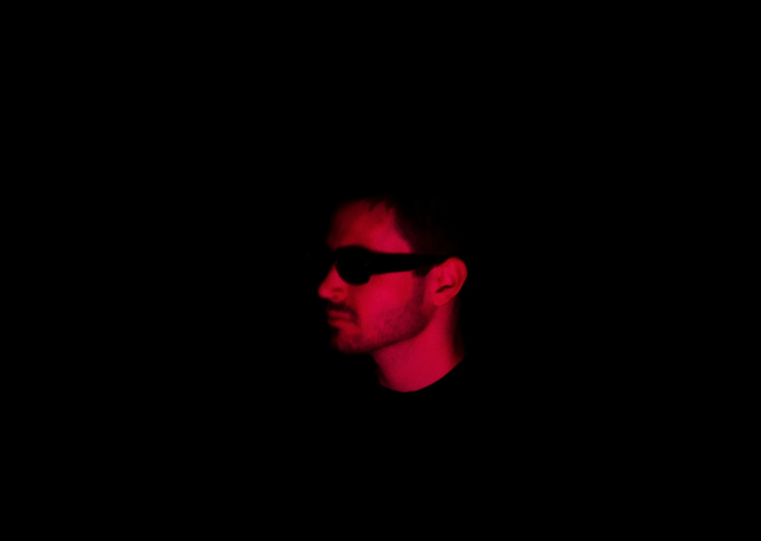 Side profile of a man wearing black sunglasses, illuminated with red lighting, against a black background.