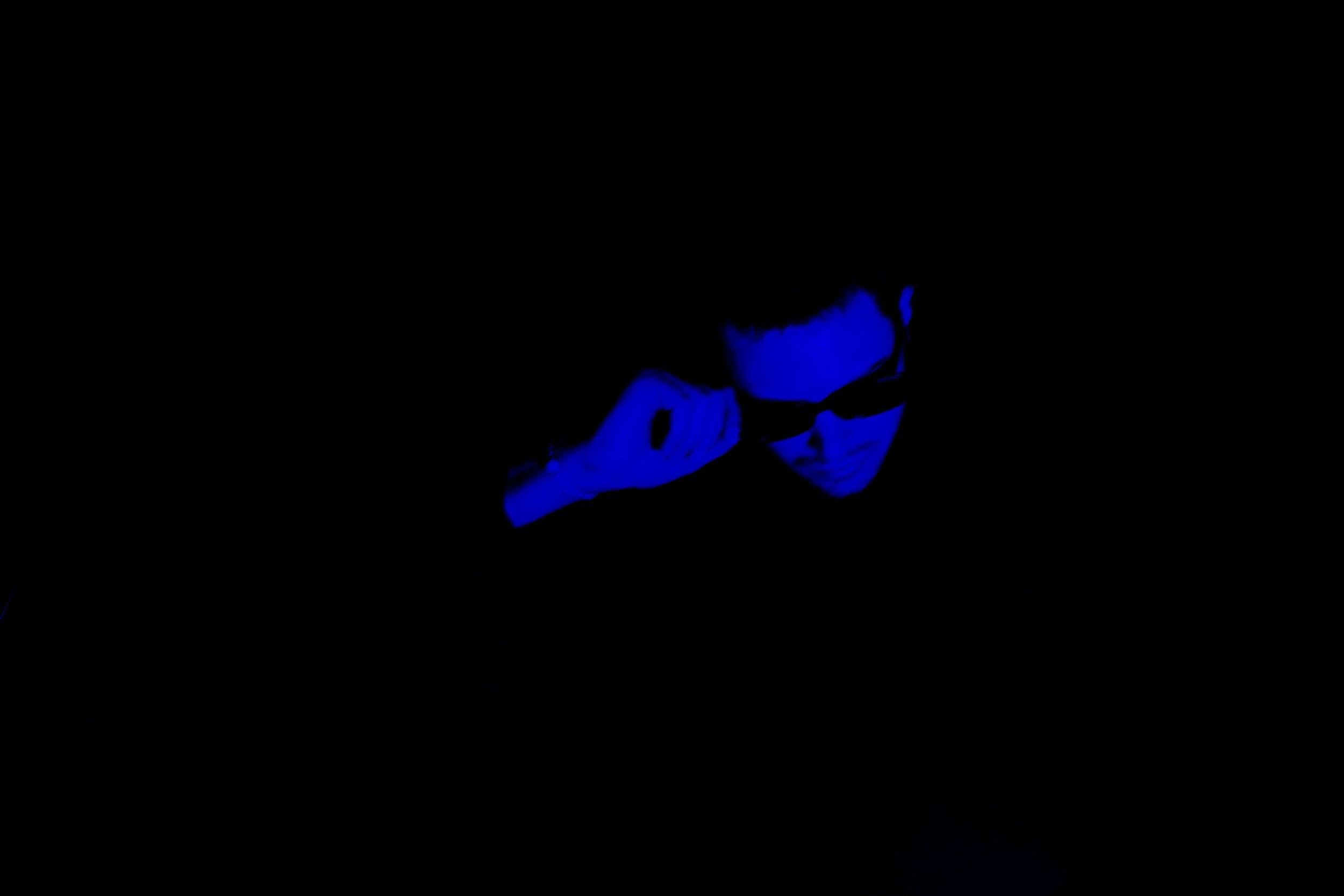 Person wearing dark sunglasses and a jacket, illuminated by blue lighting, adjusting their sunglasses in a dark environment.